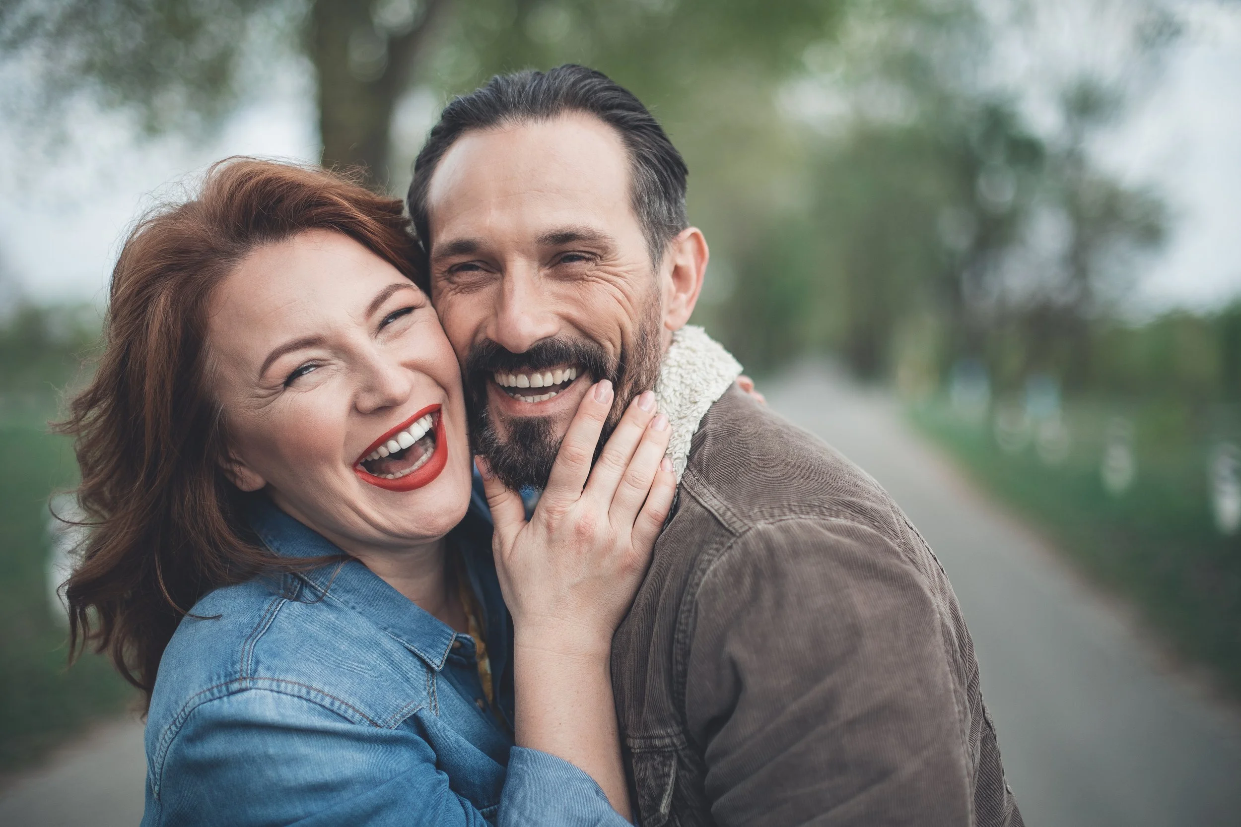 A woman and man smiling and laughing while hugging outdoors with trees and a pathway in the background.