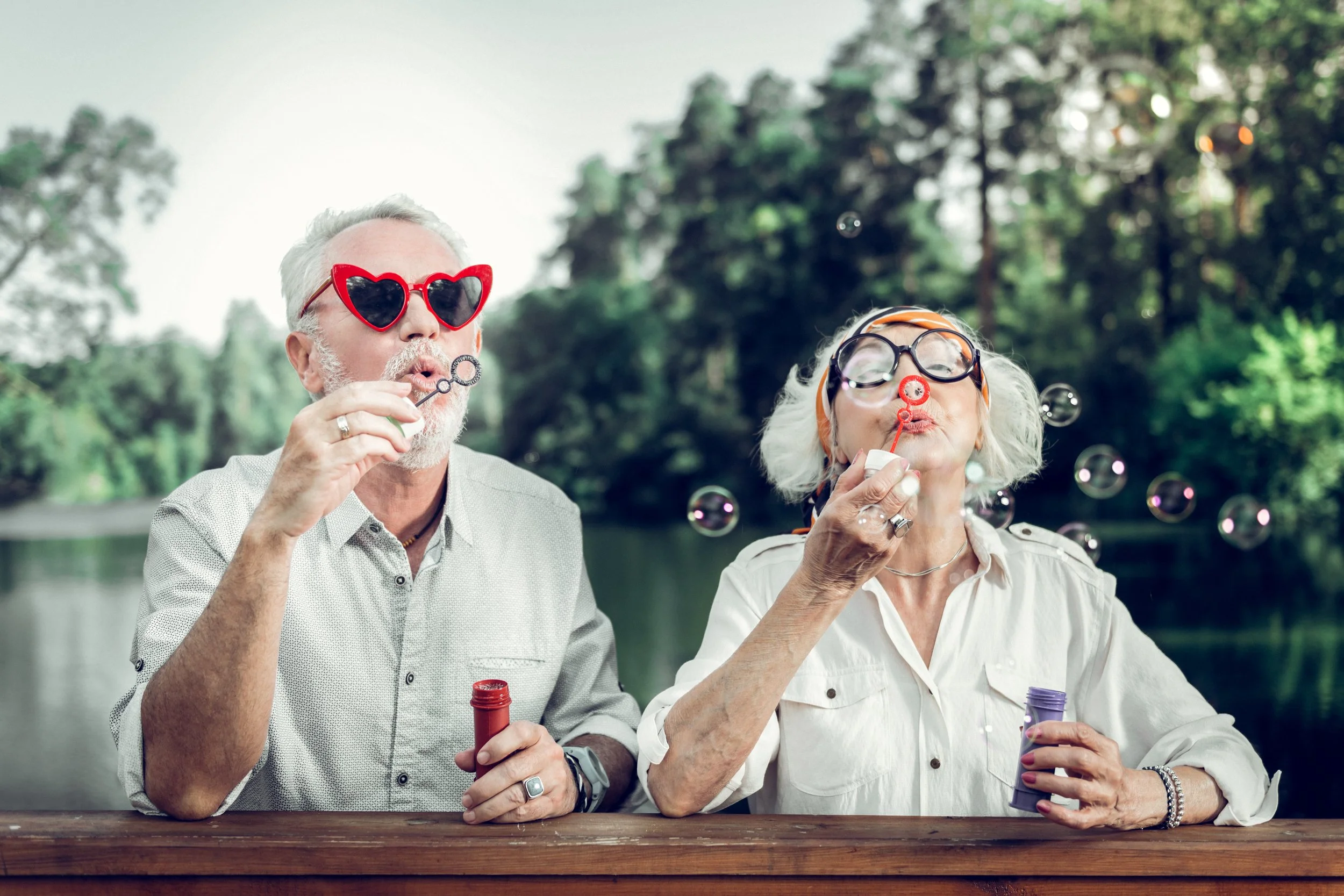 And older couple dressed in funny glasses and blowing bubbles at a picnic table outside
