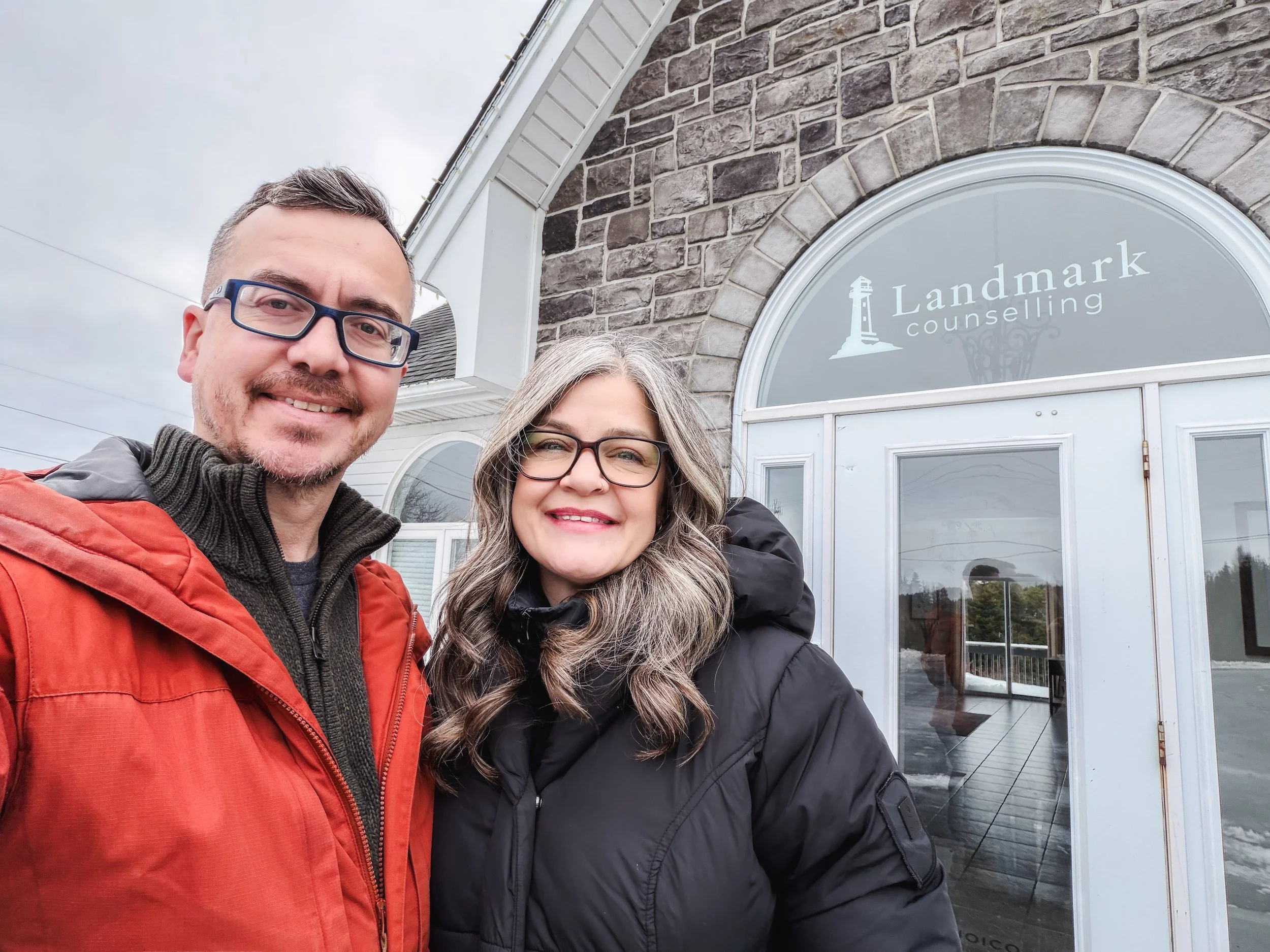 A smiling man and woman taking a selfie outside of a building with a sign reading 'Landmark counseling.' The man is wearing glasses and a red jacket, and the woman has long, wavy hair and is wearing glasses and a black jacket.