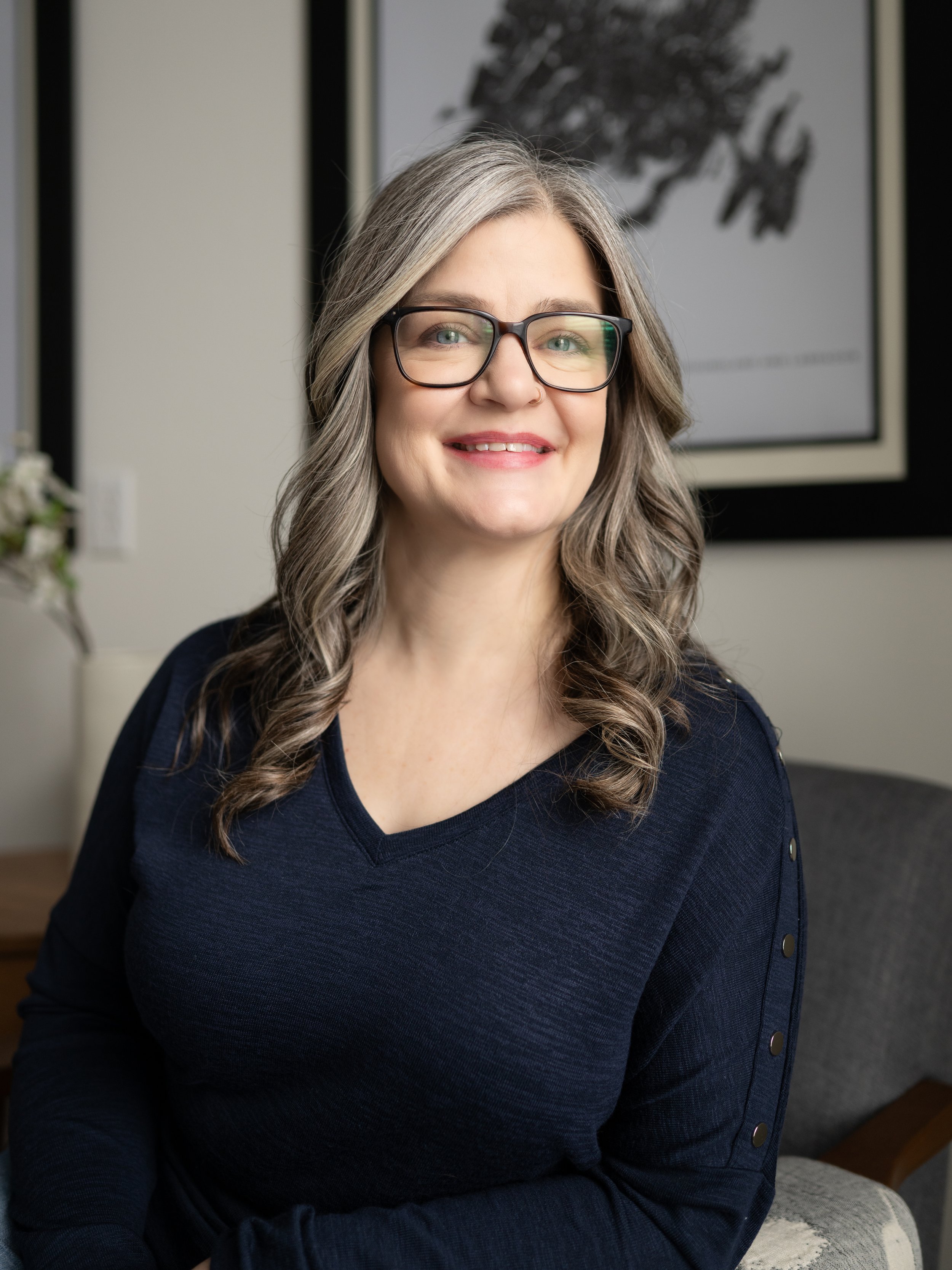 A woman with long gray hair wearing glasses and a navy blue top, smiling and sitting in a modern indoor setting.