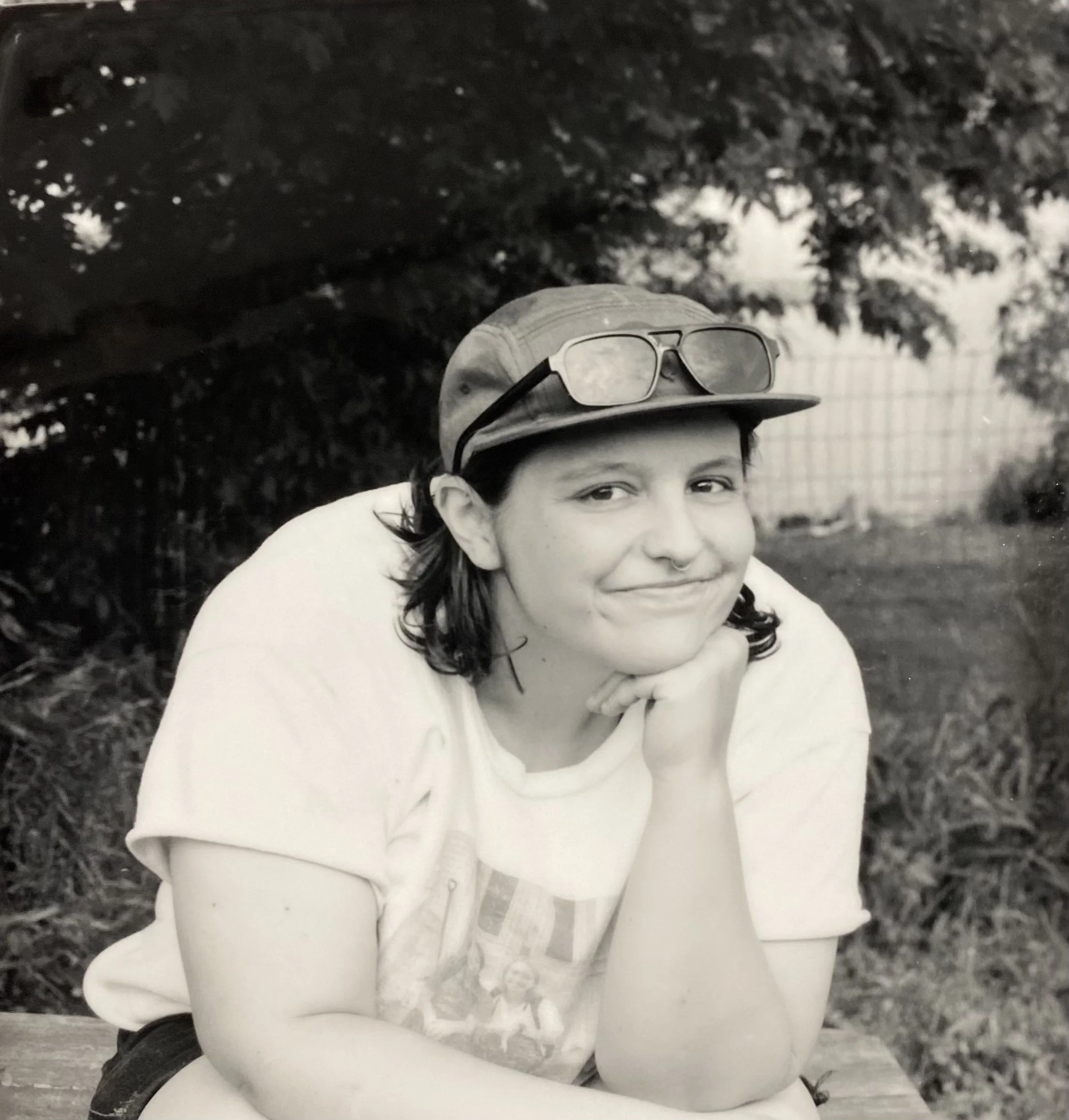 A young person with shoulder-length dark hair, wearing a cap with sunglasses on top, smiling and resting their chin on their hand outdoors.