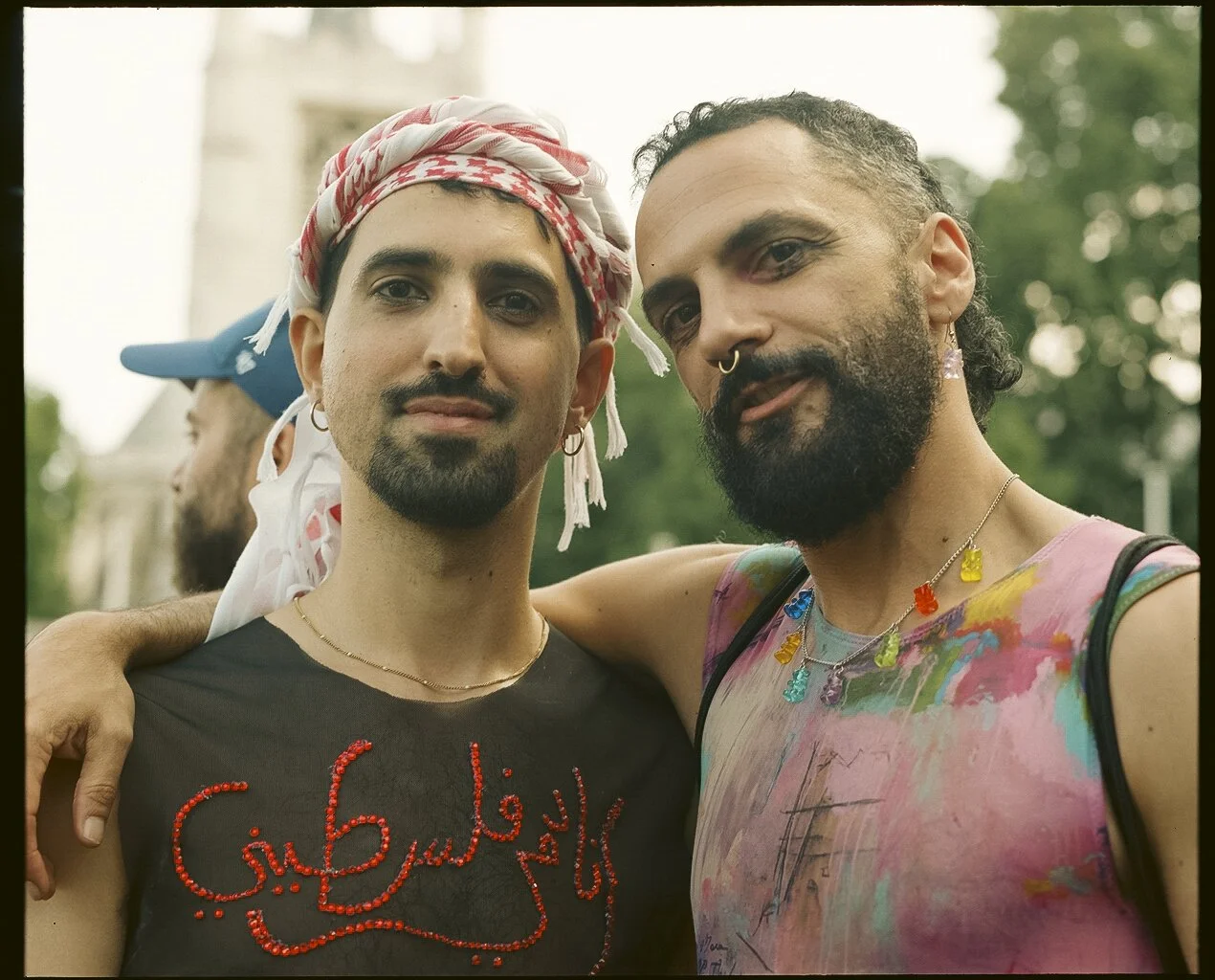 Close-up portrait of two people. The person on the left wears a white and red keffiyeh and a black tank top with a red design, while the person on the right wears a colourful paint-splatter tank top and has their arm around the other’s shoulders. The