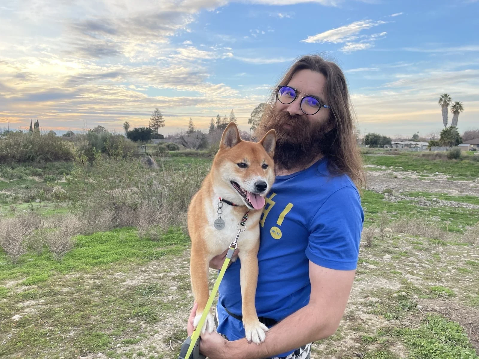 A man with long hair, glasses, and a beard holding a Shiba Inu dog outdoors during sunset, with a landscape of grass, trees, and a partly cloudy sky in the background.