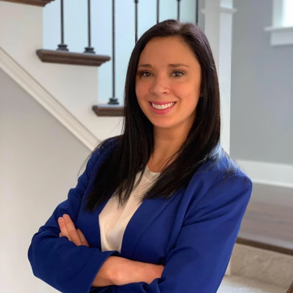A woman with long dark hair, wearing a blue blazer over a white top, smiling with her arms crossed, standing indoors near a staircase with black railings.