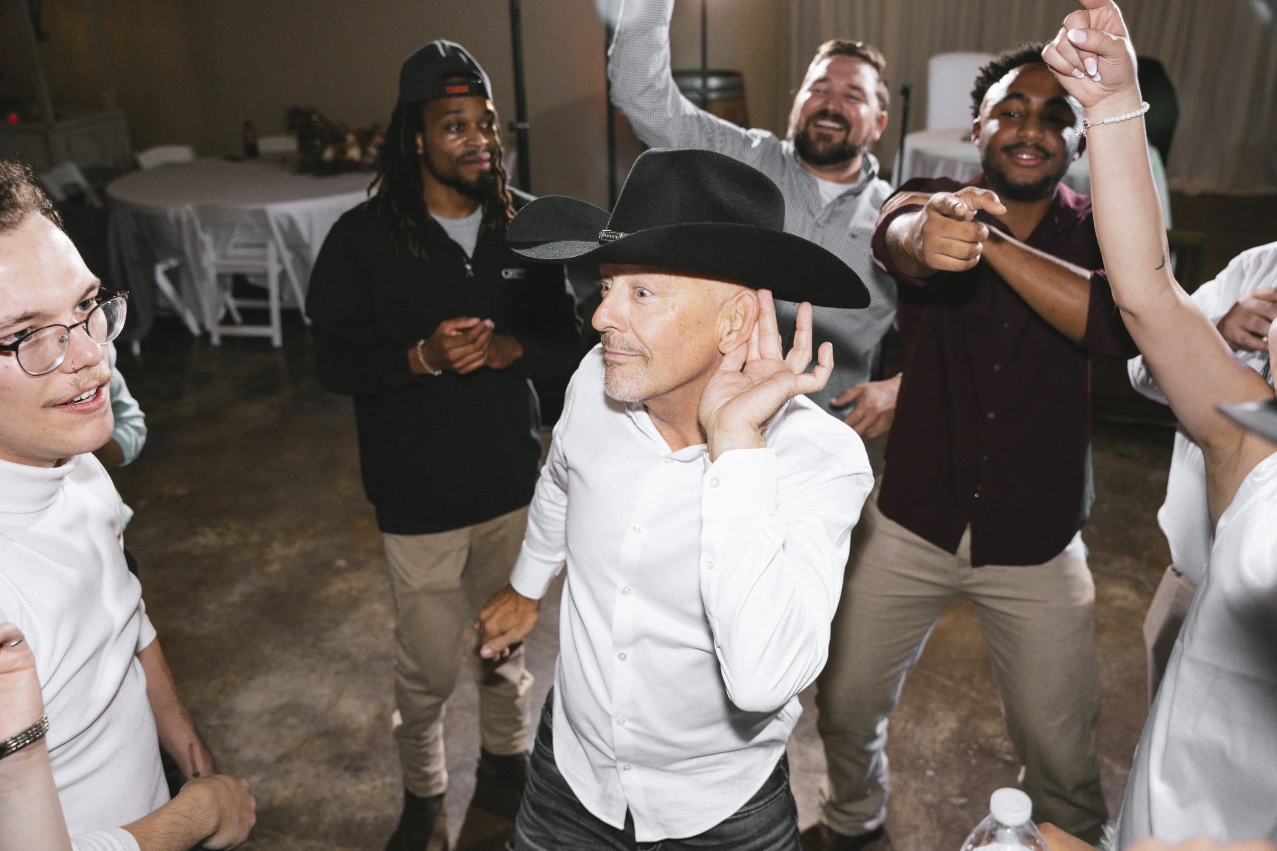 A man wearing a black cowboy hat and white shirt is dancing at a social event, surrounded by several other people who are also dancing and having fun.