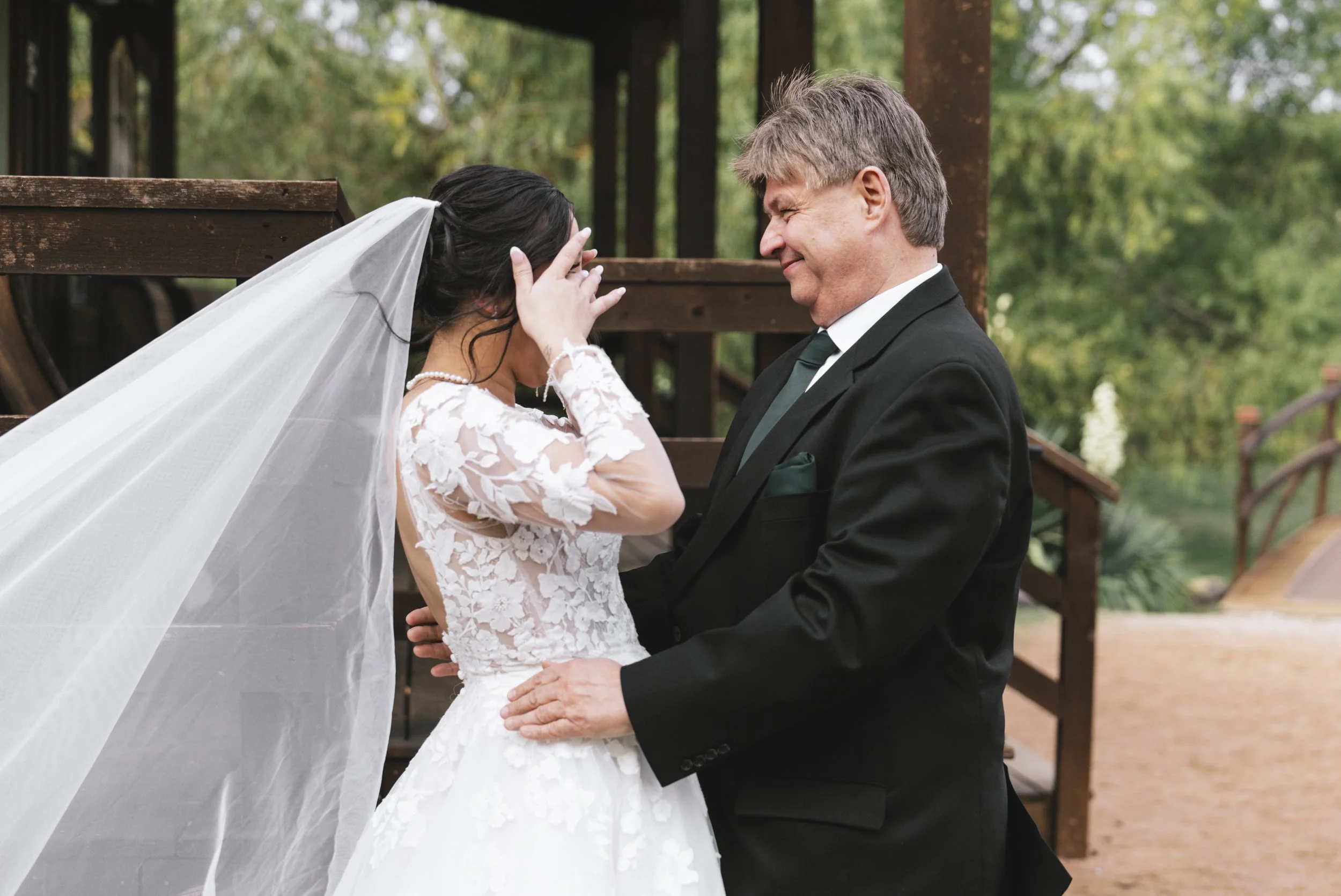 A bride and her father share a heartfelt moment on her wedding day, standing outdoors on a wooden bridge, with greenery in the background.