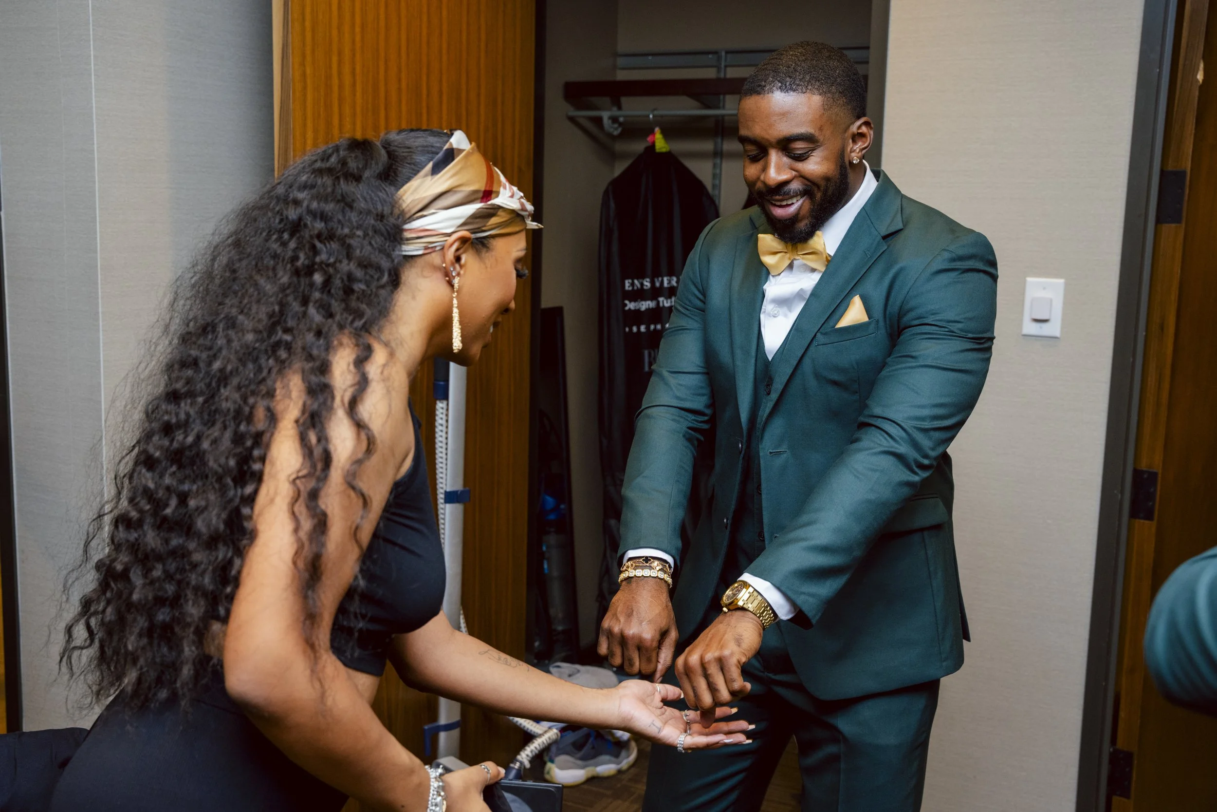 A man in a teal suit and gold bow tie and a woman in a black dress are laughing and showing their rings together in a closet. The man is wearing a gold watch, and the woman is wearing earrings and a scarf in her hair.
