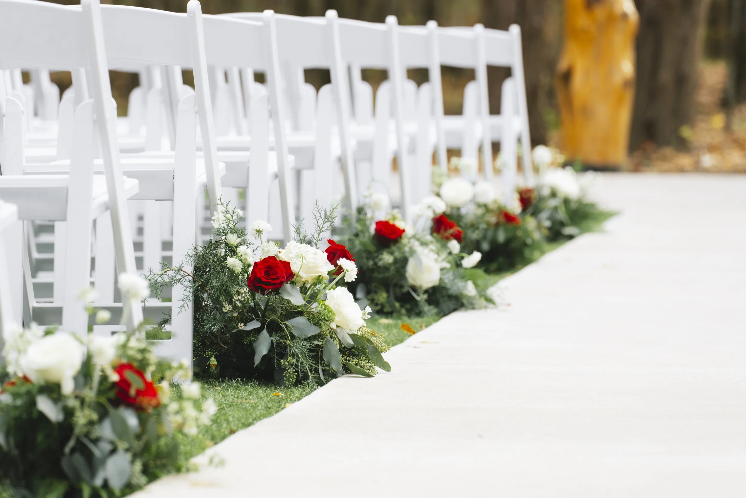 Row of white folding chairs decorated with white and red flowers along a white aisle at an outdoor wedding ceremony site.