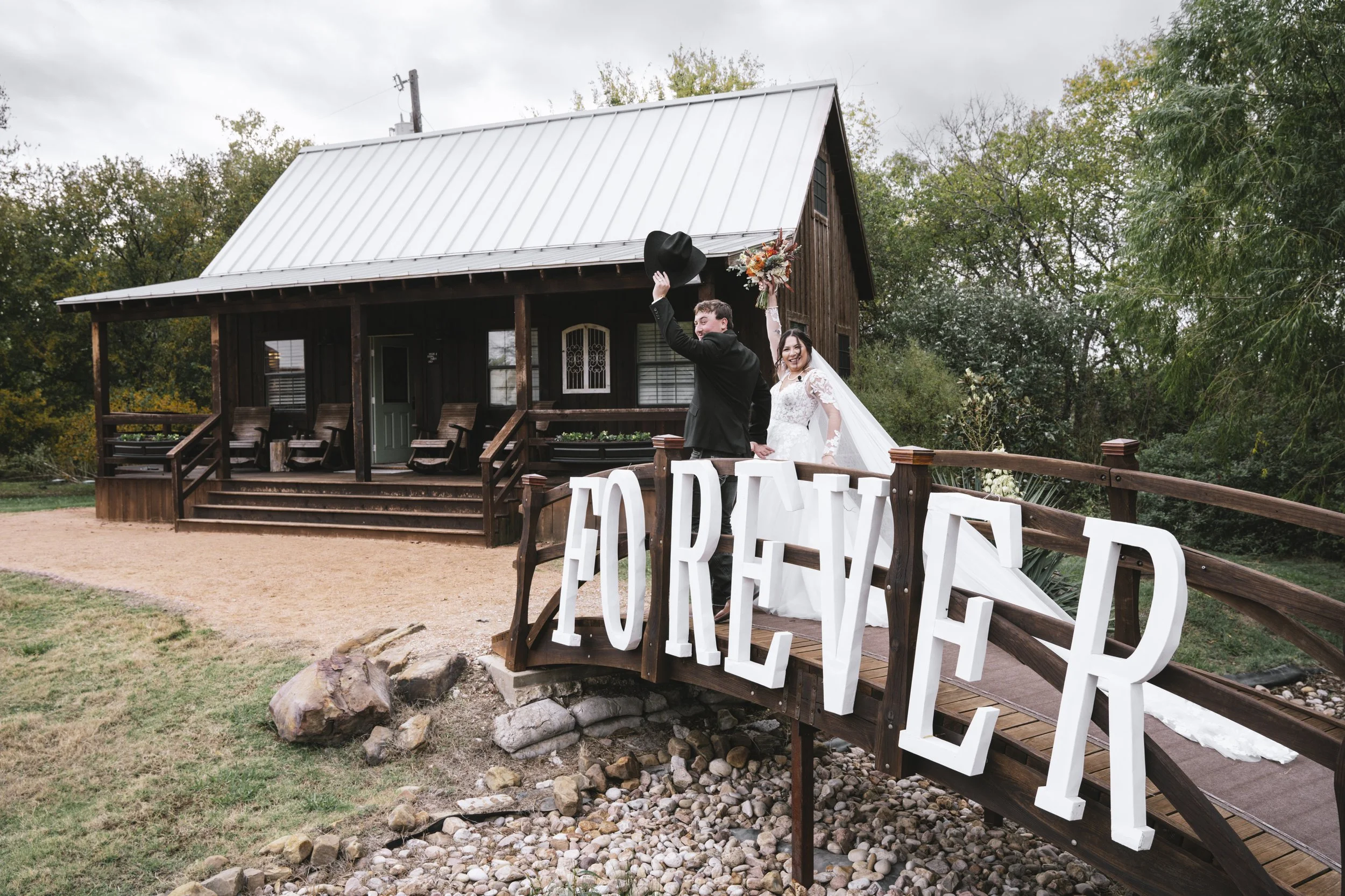 A newlywed couple celebrating on a wooden bridge with a large sign that reads 'FOREVER' in front of a rustic house surrounded by trees.