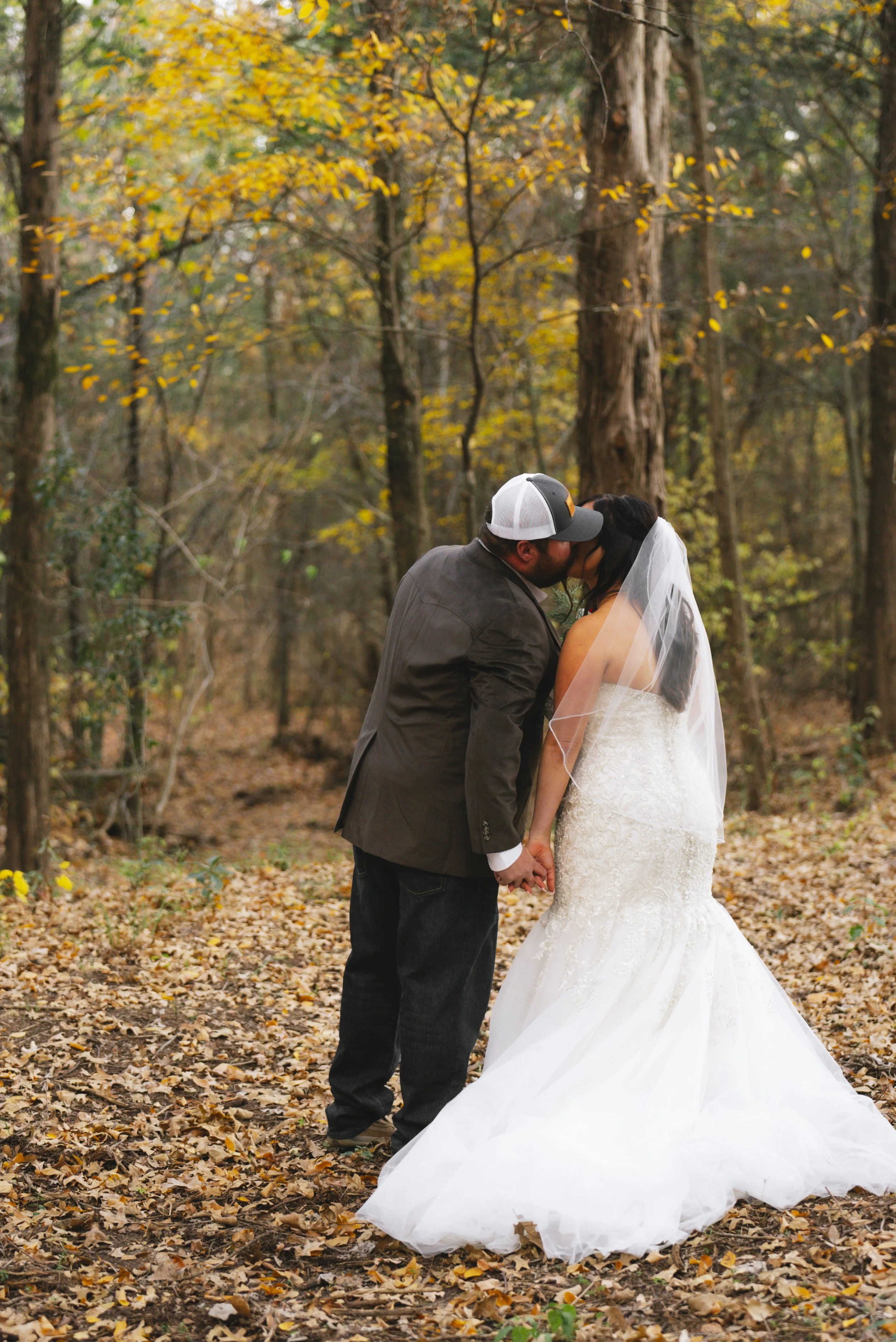 A couple in wedding attire kissing in a forest with fallen autumn leaves.