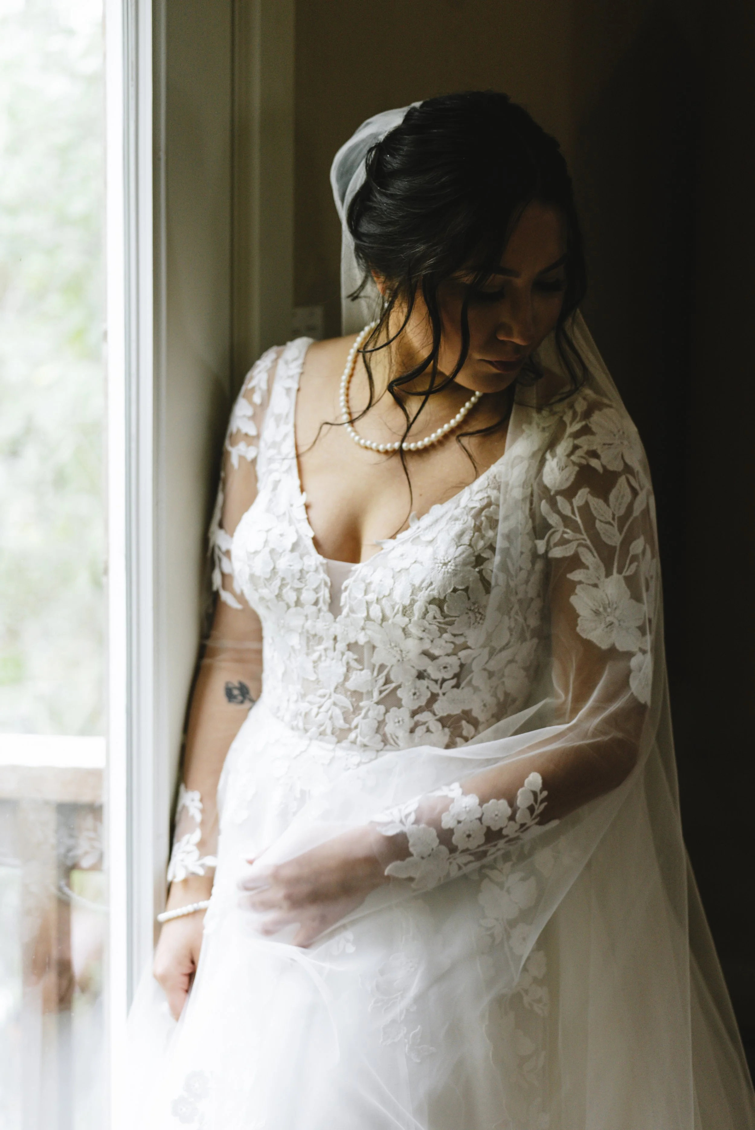 Bride in a white lace wedding dress with floral patterns, standing by a window, looking down, with dark, wavy hair and a pearl necklace.
