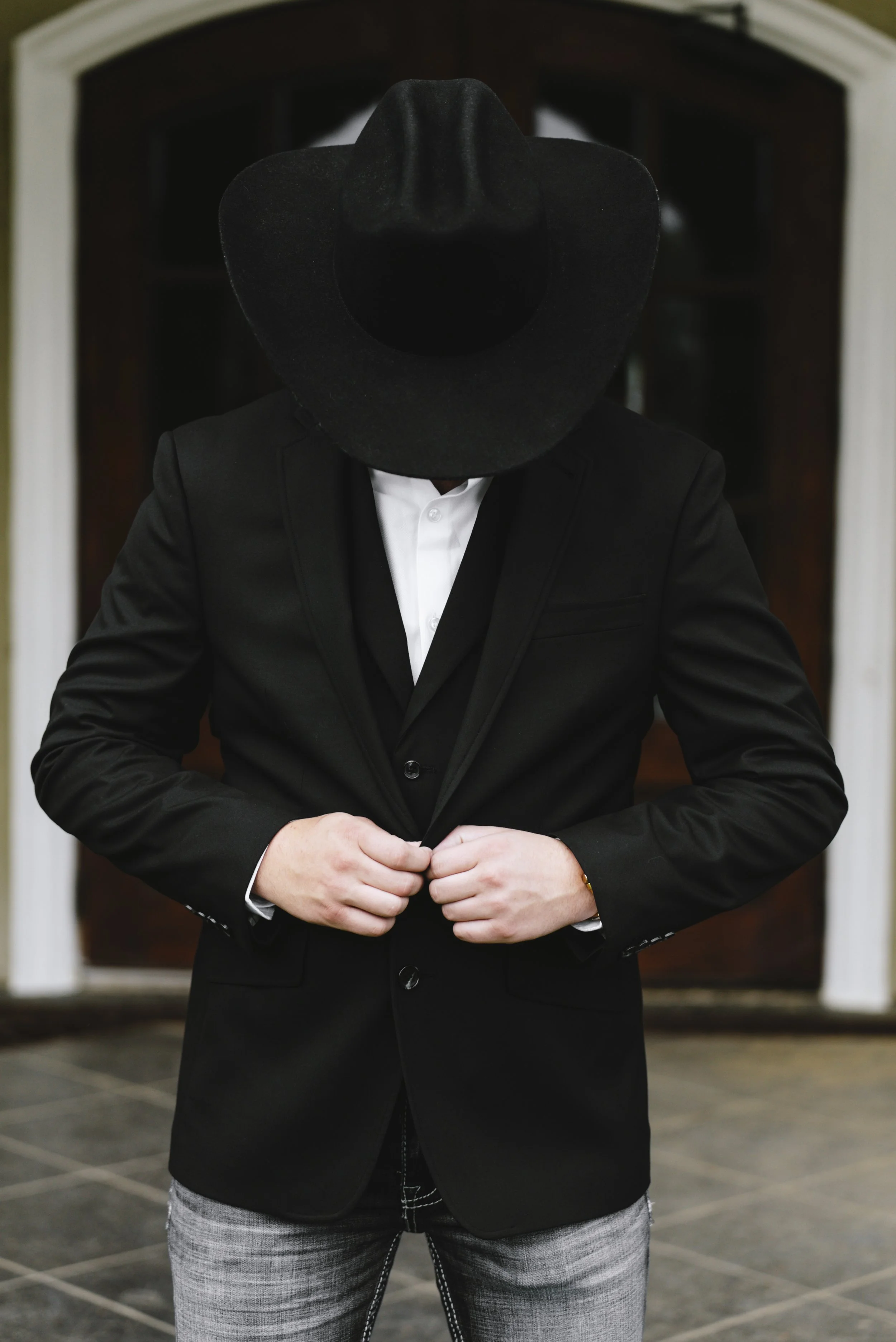 Person wearing a black blazer, white shirt, and a large black cowboy hat, standing outdoors in front of a wooden door.