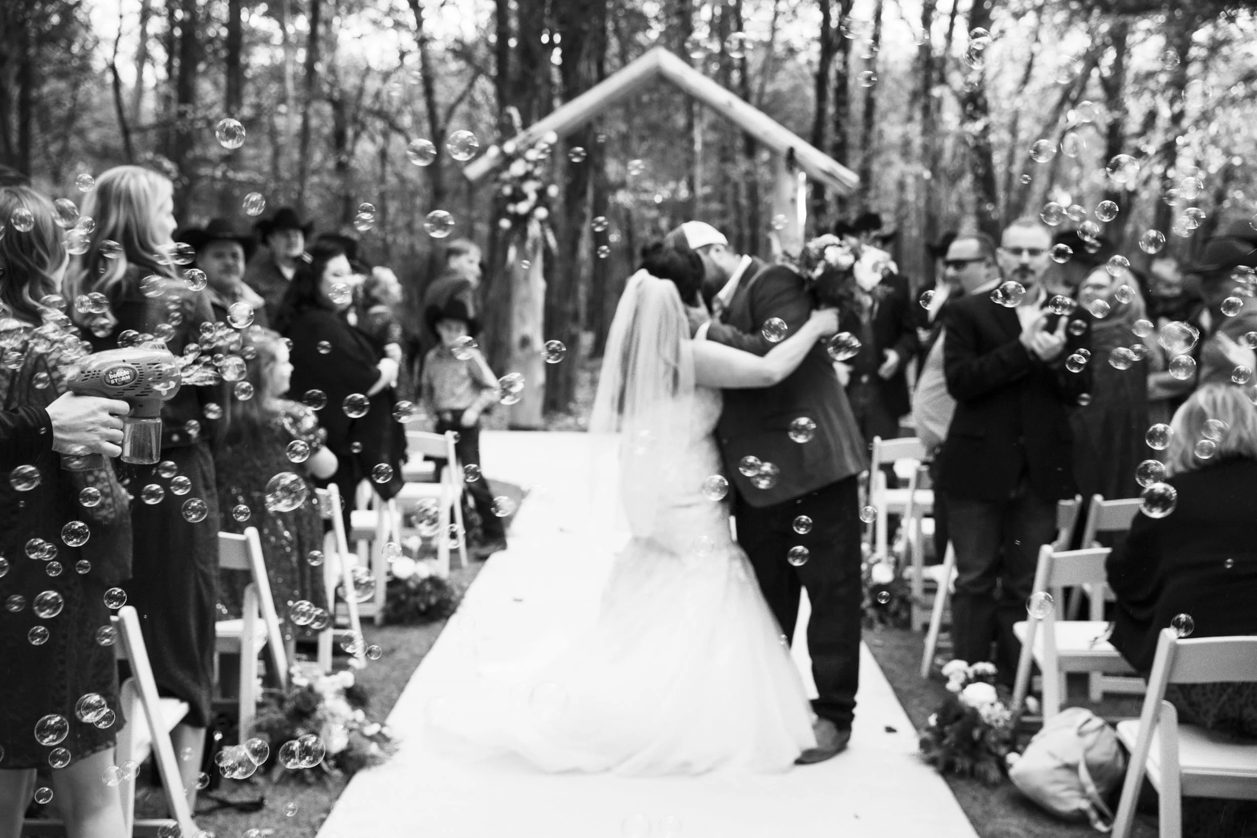 A black and white photo of a wedding ceremony in a wooded outdoor setting, with a couple kissing at the altar, surrounded by guests and bubbles.