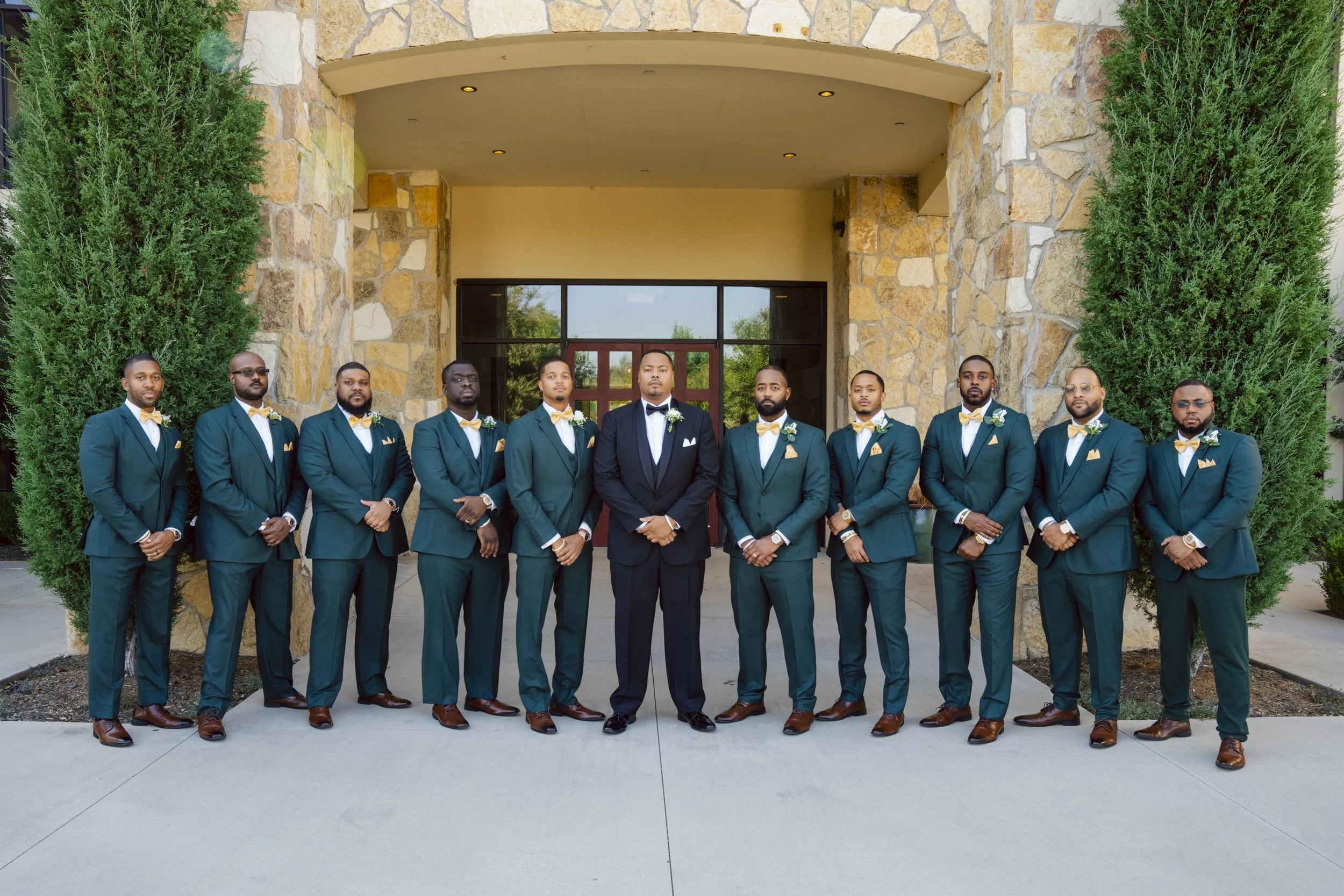 A groom in a black suit and ten groomsmen in matching teal suits with yellow bow ties, all with white pocket squares and boutonnieres, standing in a line outside a stone building with large green trees on either side.