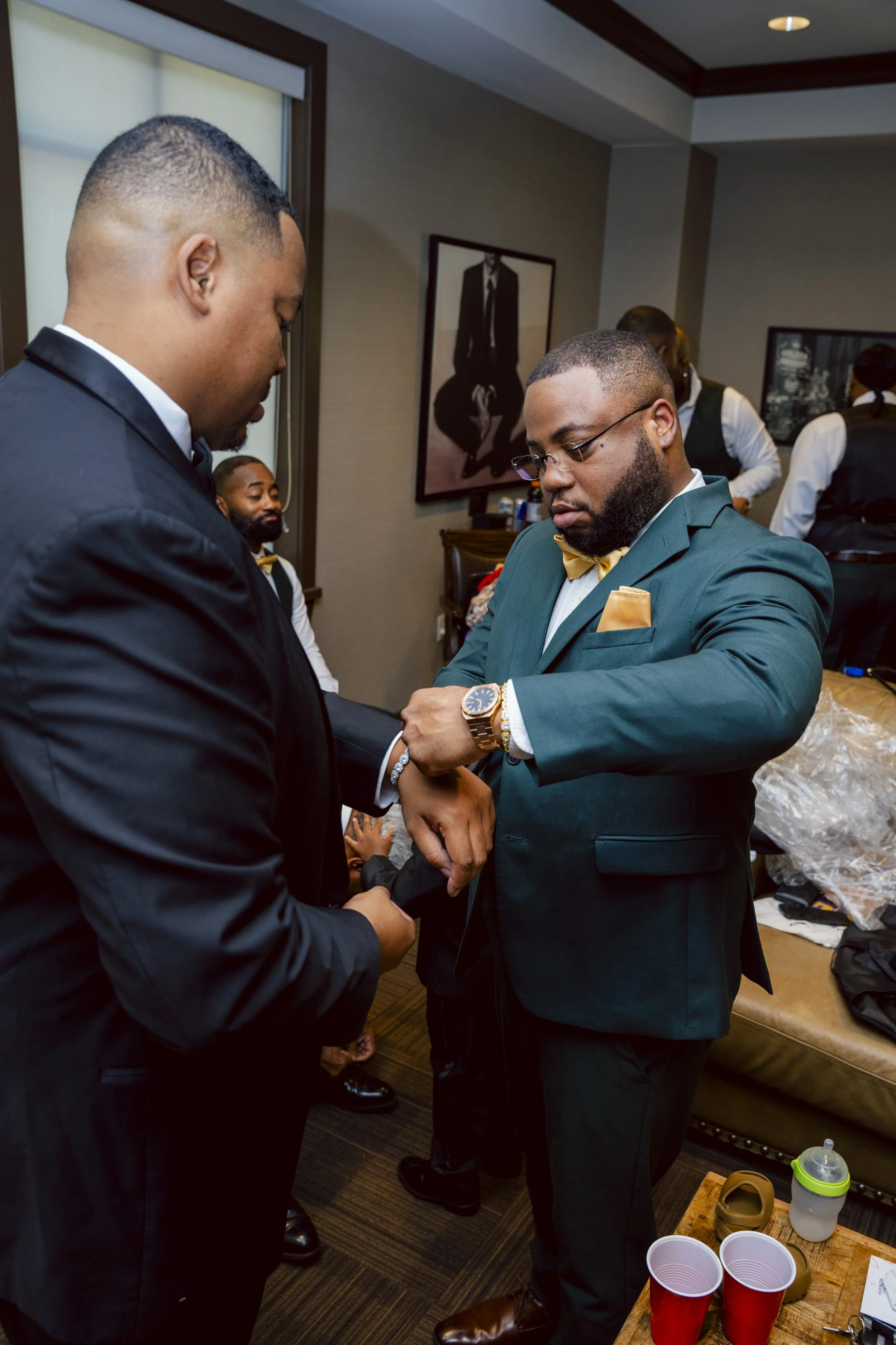 Two men in formal suits, one with glasses, are adjusting his cufflink in a room with other people and artwork on the wall, preparing for a formal event.