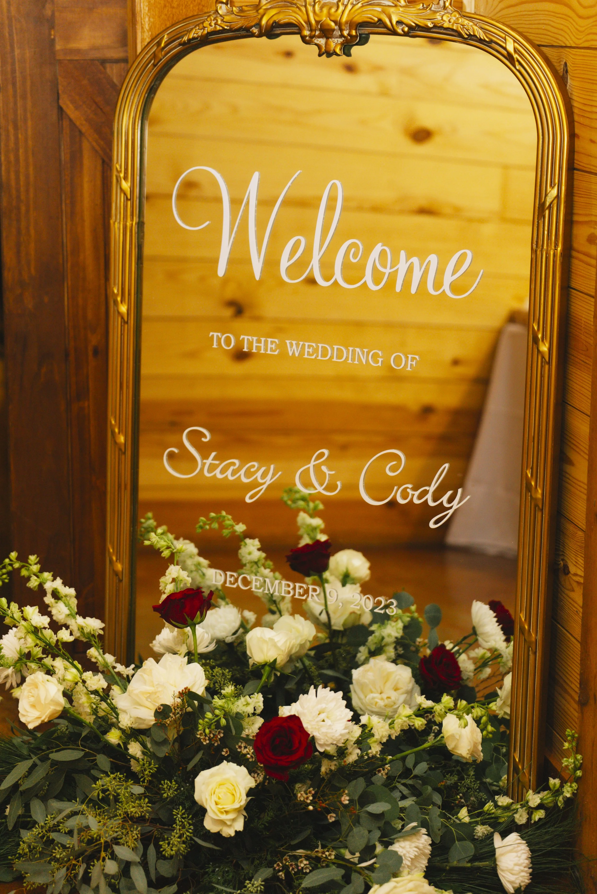 Decorative mirror with wedding welcome sign for Stacy and Cody, dated December 9, 2023, surrounded by white and dark red flowers and greenery.