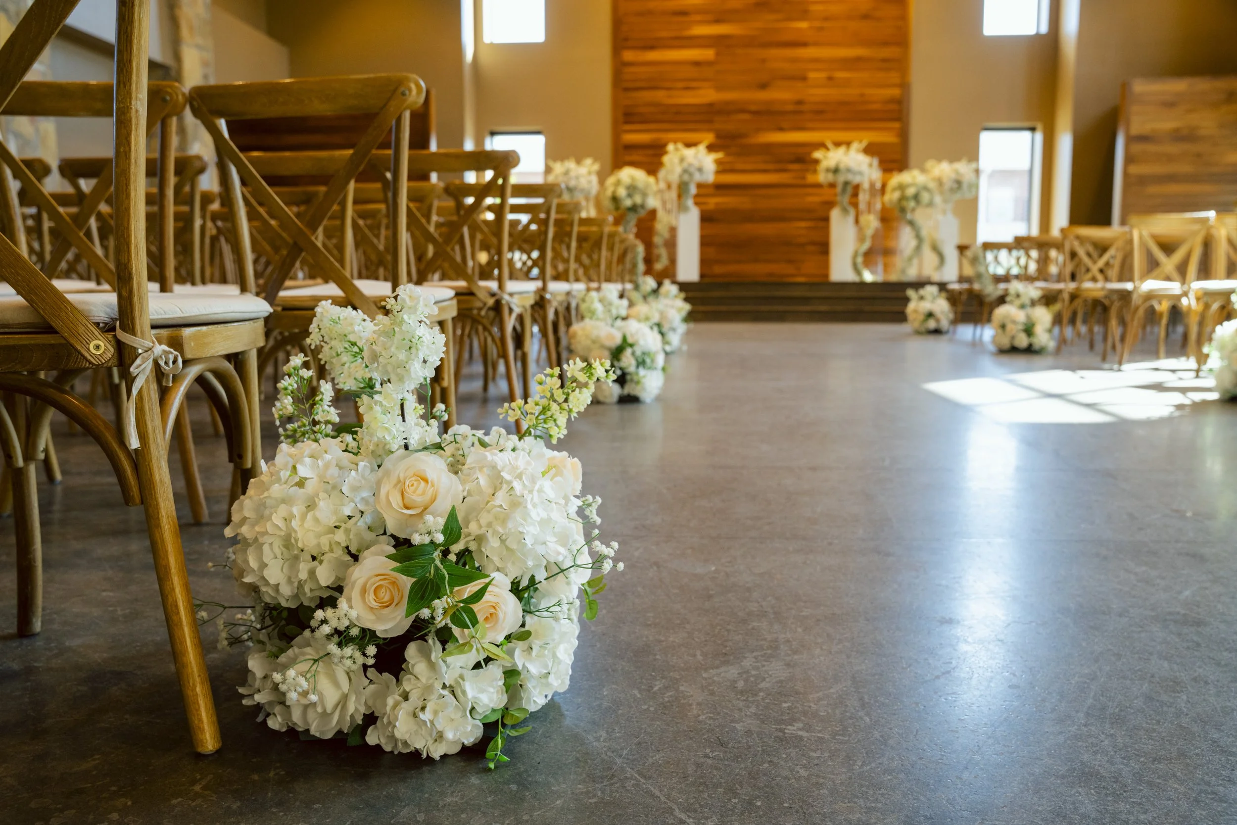 Wedding ceremony decor with white floral arrangements lining the aisle, multiple wooden chairs on either side, and an elevated altar at the front in a warmly lit indoor setting.