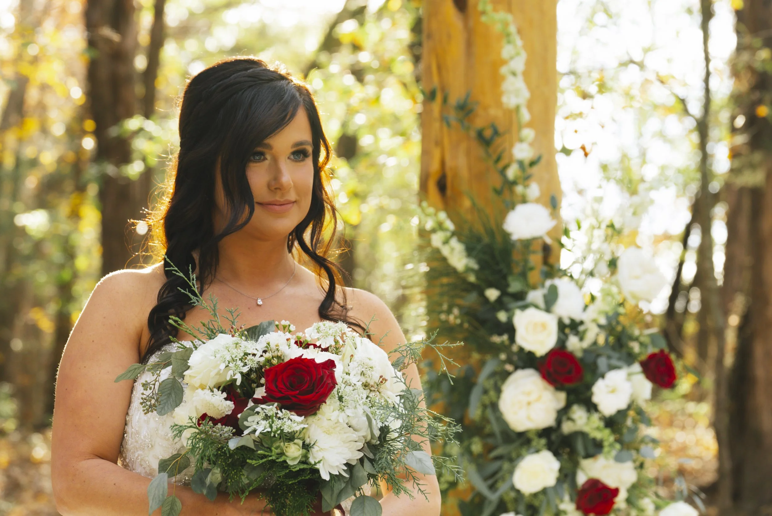 A bride with dark hair in loose waves holding a bouquet of white and red roses, standing outdoors near floral decorations in a wooded area.