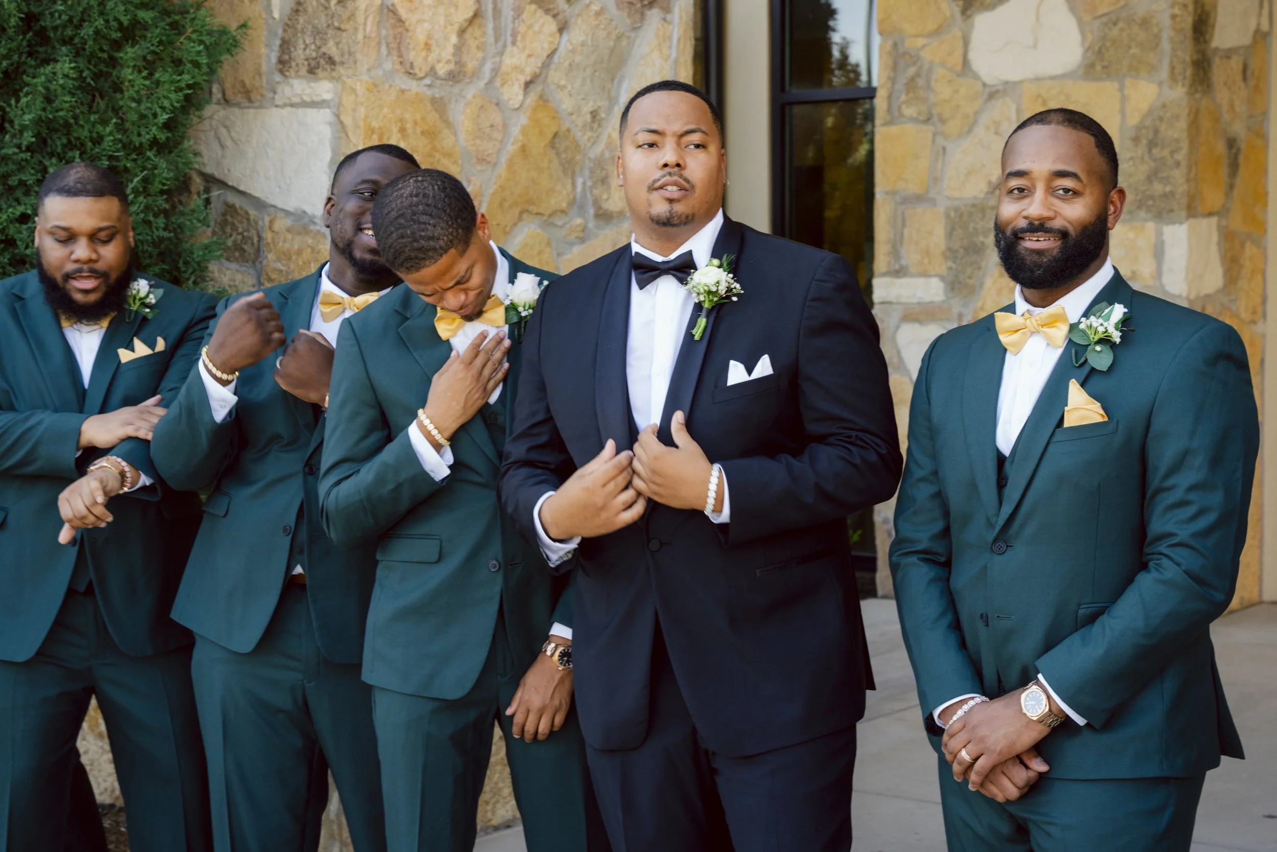 A group of five men in tuxedos, with one man adjusting his suit, standing outside near a stone wall. Four men are wearing dark green tuxedos with yellow bow ties, and one man in the middle is wearing a black tuxedo with a black bow tie. They are all 