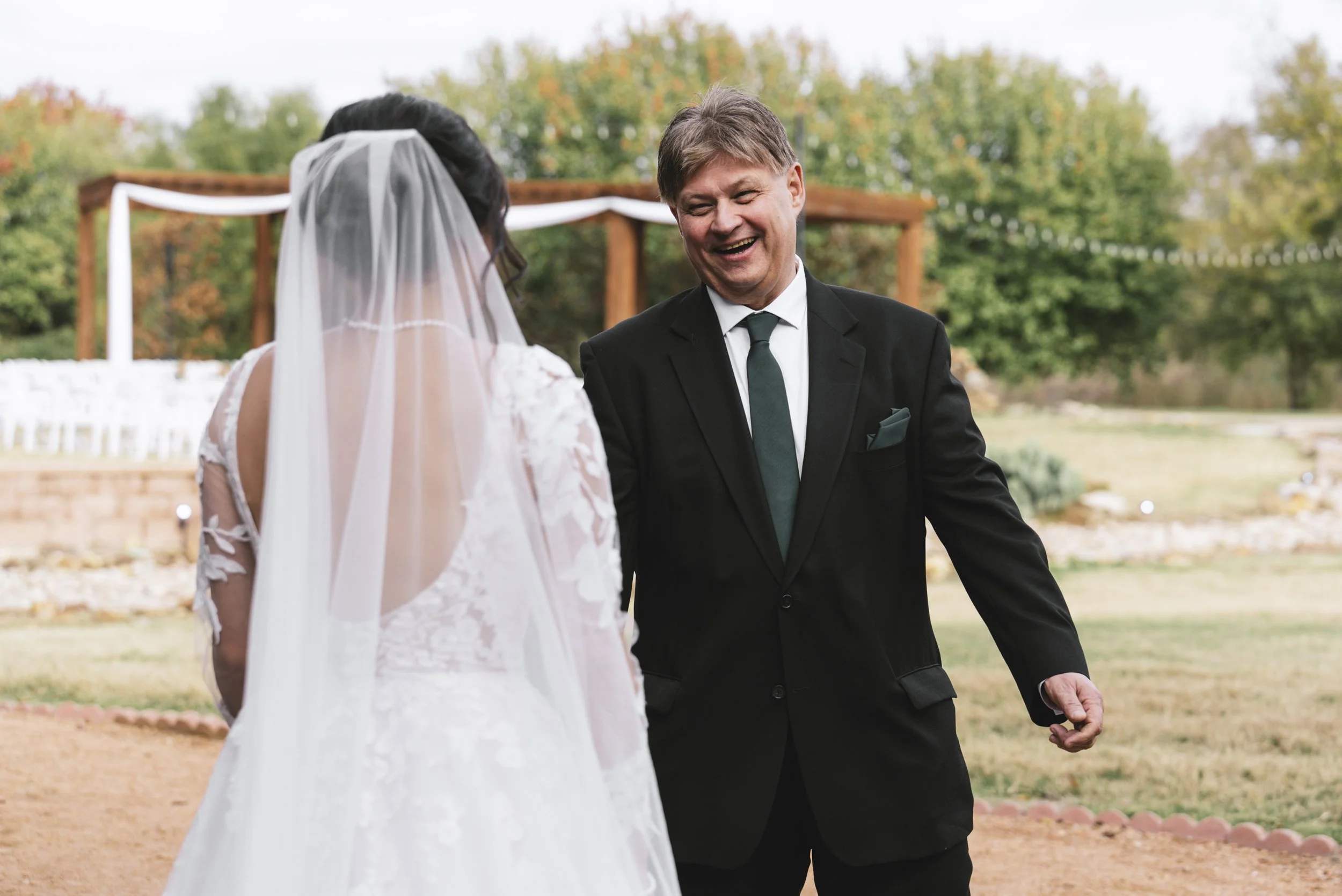 A bride and a man, likely her father, smiling and talking outdoors during a wedding ceremony, with trees in the background.