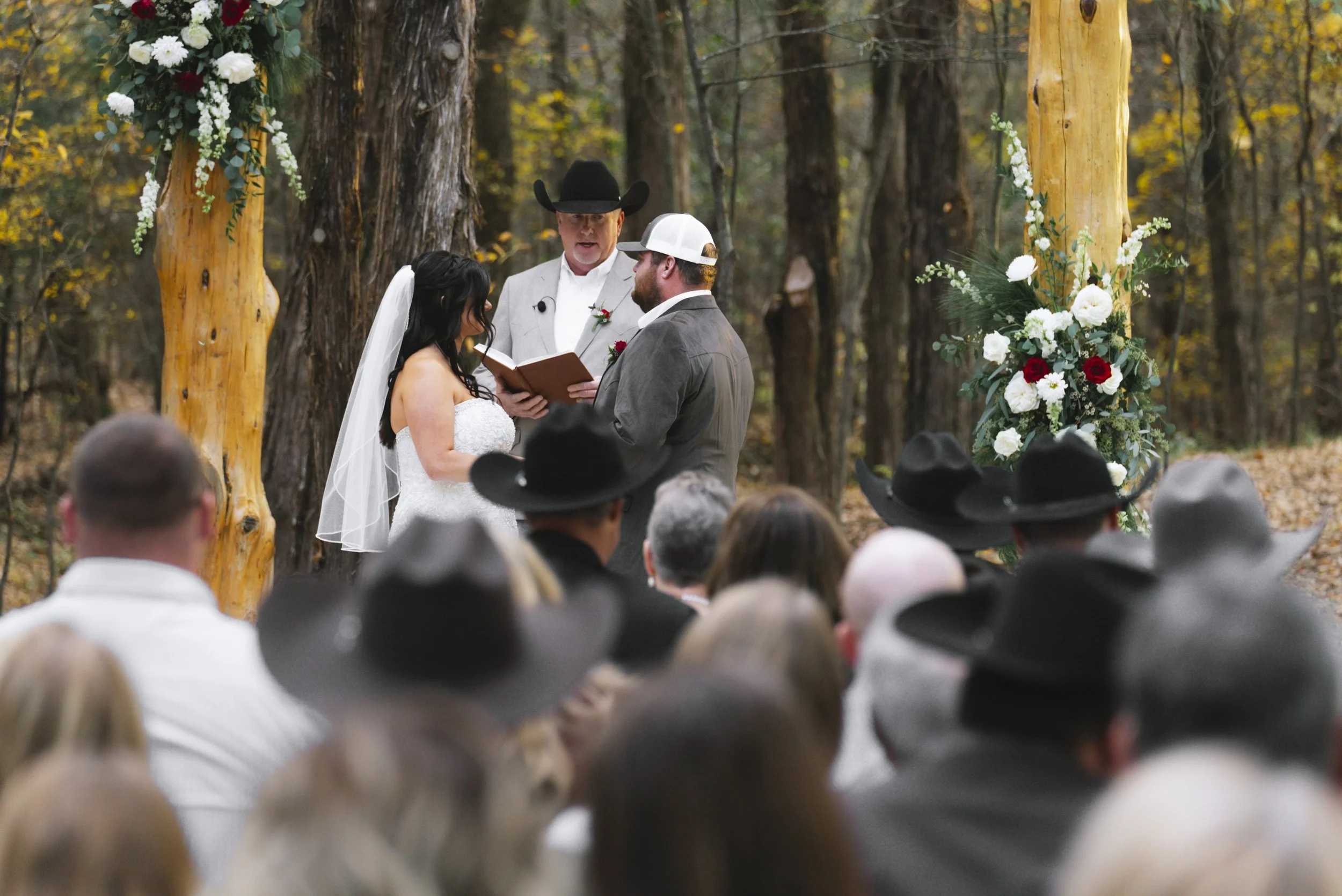 A couple getting married outdoor in a forest, exchanging vows under a decorated wooden arch, with an officiant in gray and a group of guests watching, some wearing cowboy hats.