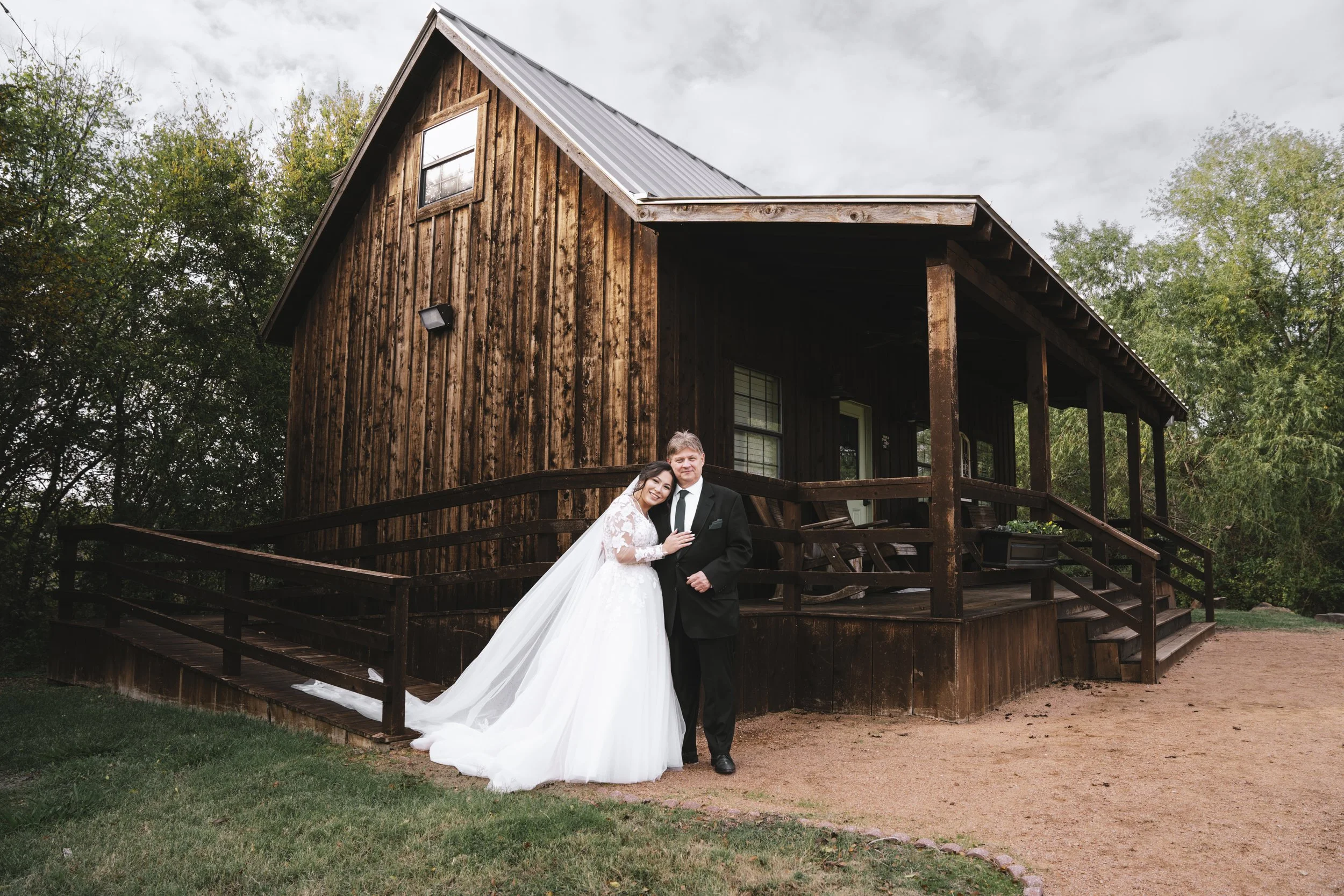 Bride and groom in wedding attire standing in front of a wooden house with a porch and steps, surrounded by trees.