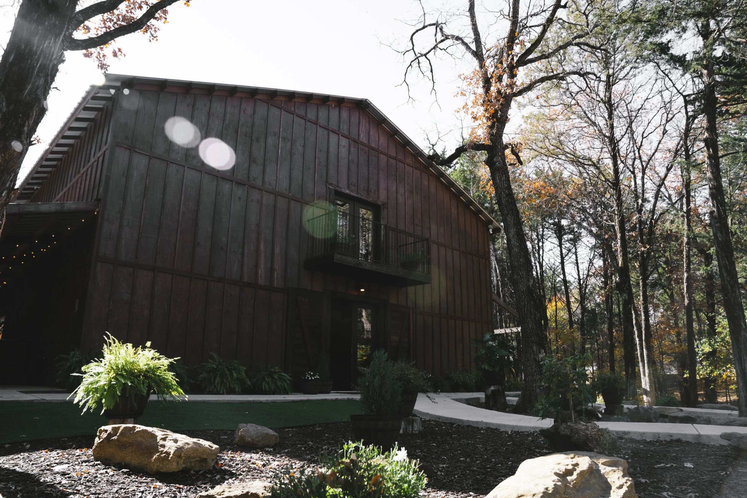A rustic wooden house surrounded by trees on a sunny day with some leaf fall, potted plants, large rocks, and a winding pathway.