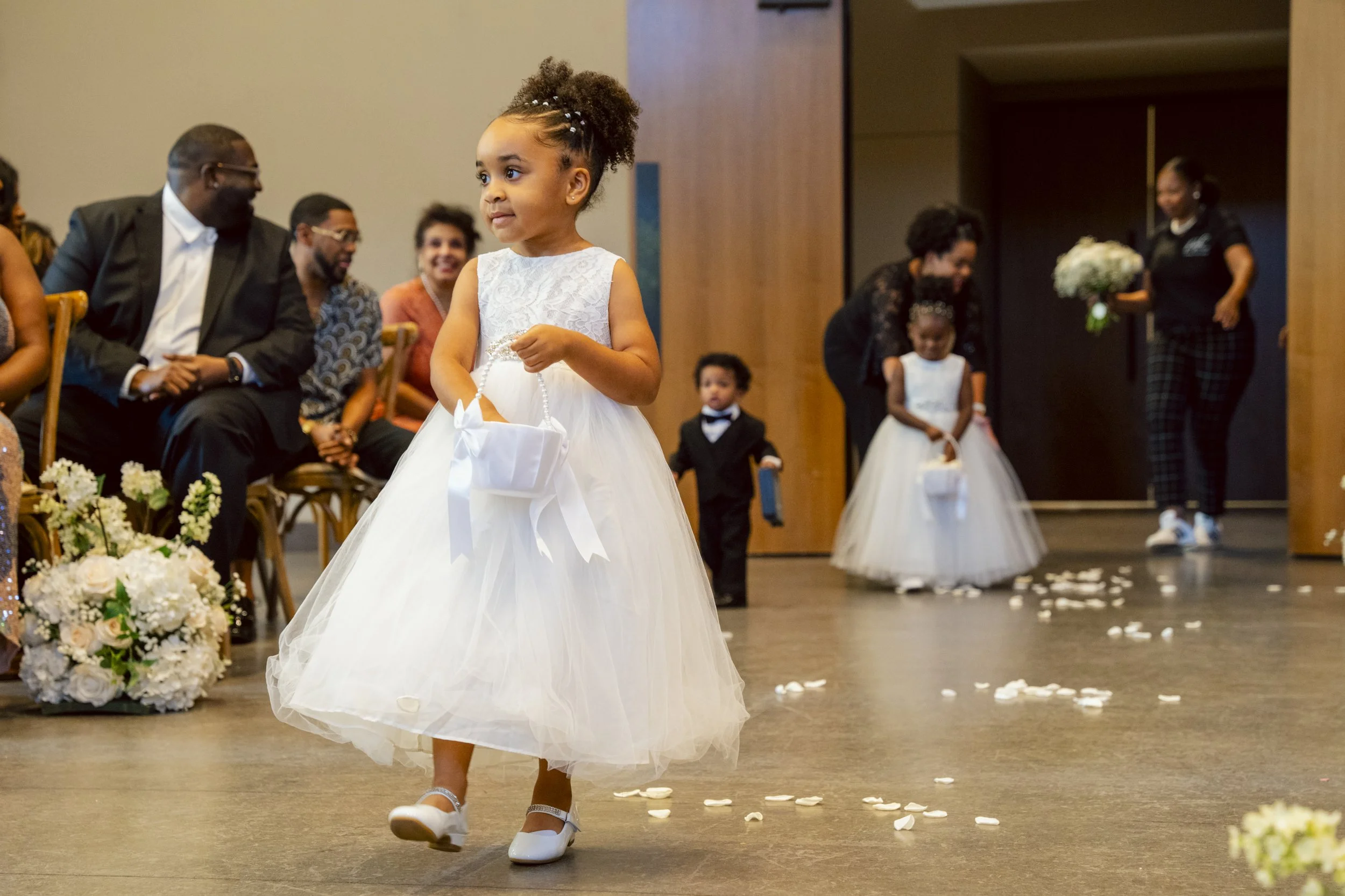 Young girl in a white dress walking down the aisle at a wedding ceremony, holding a white basket, with guests seated and smiling in the background.