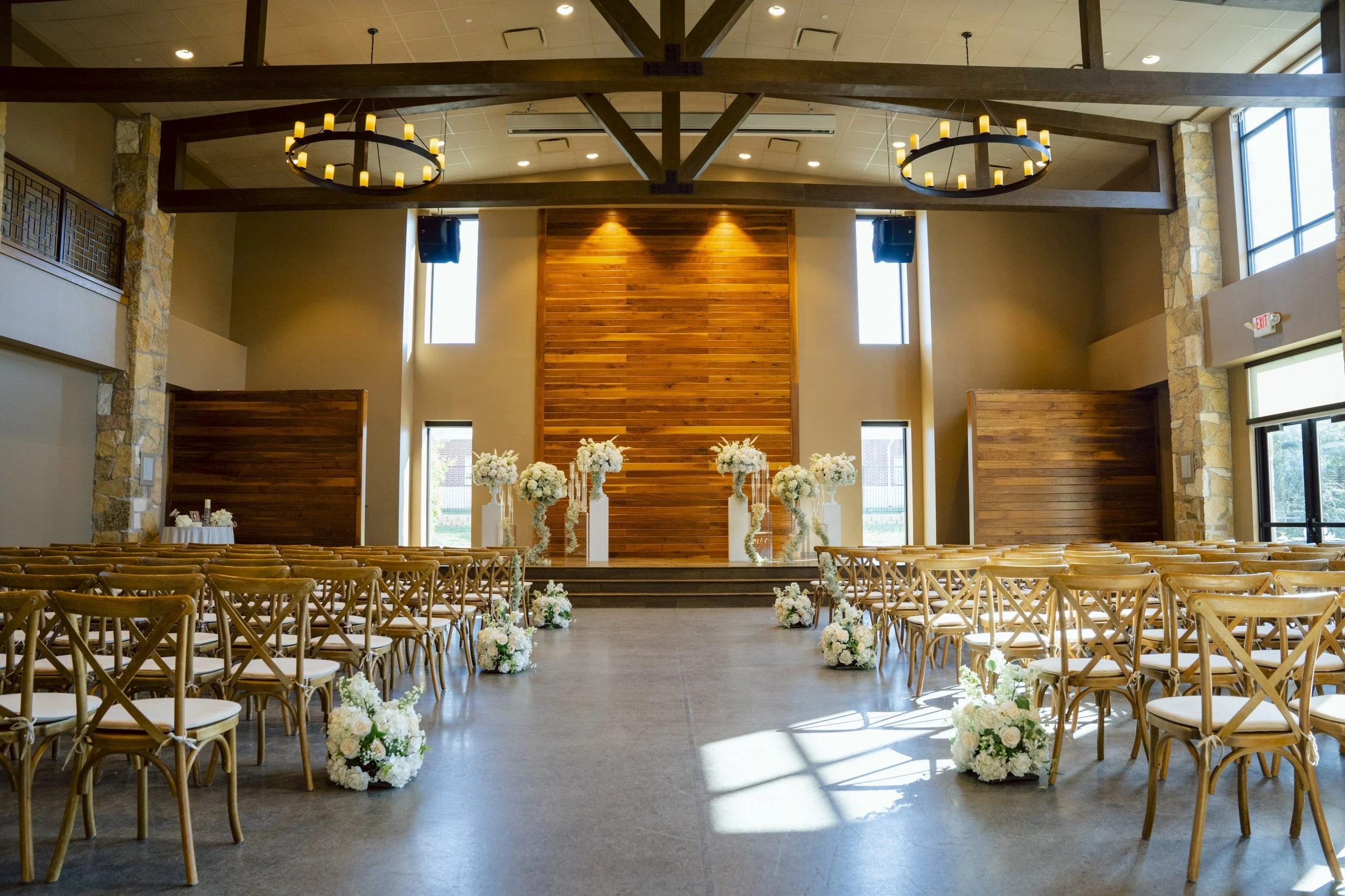Indoor wedding venue decorated with white flowers and wooden chairs, set up for a ceremony with an elevated stage and floral arrangements along the aisle.