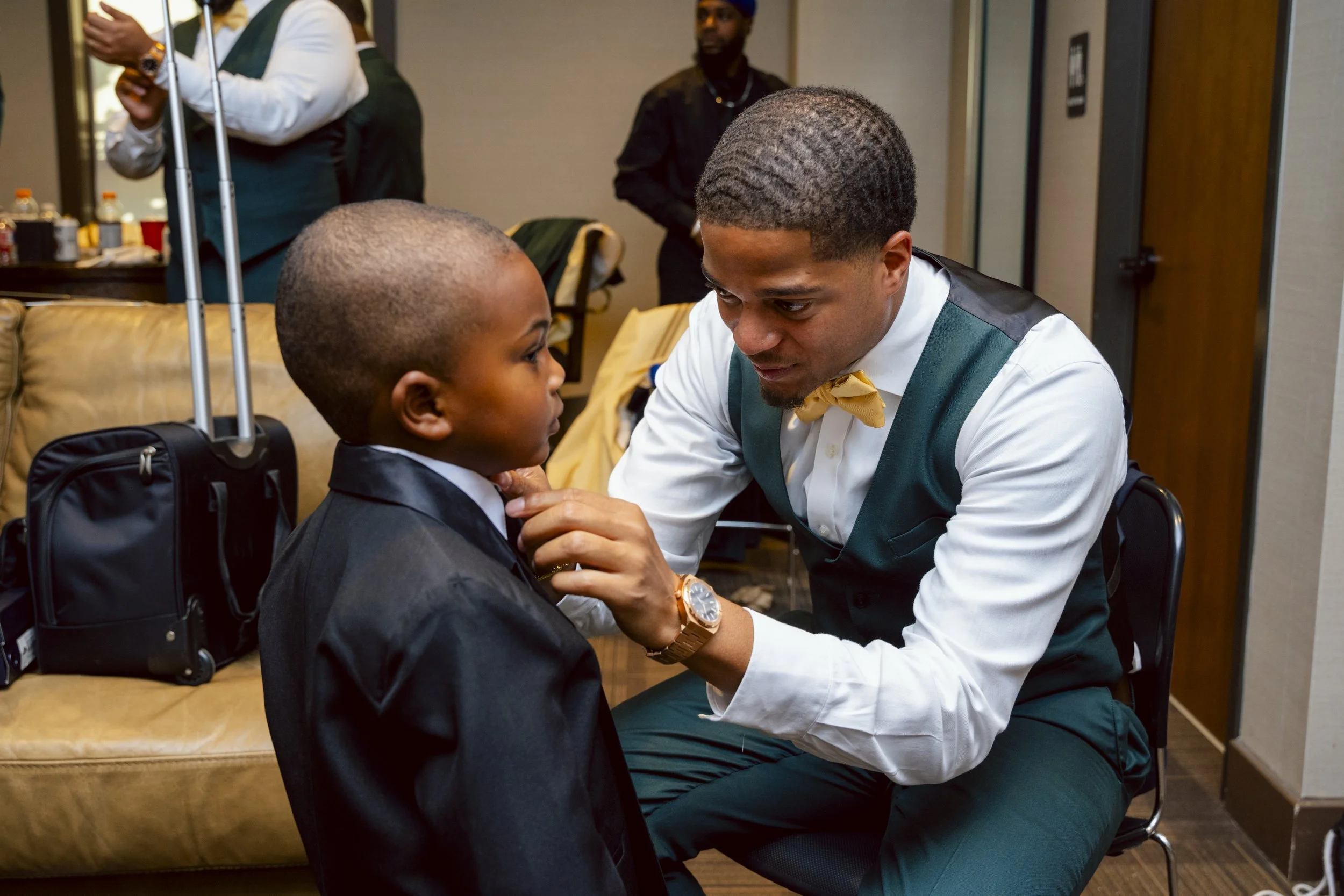A young boy in a tuxedo having his tie adjusted by an adult man in a vest with a bowtie, in a hotel room or conference setting.