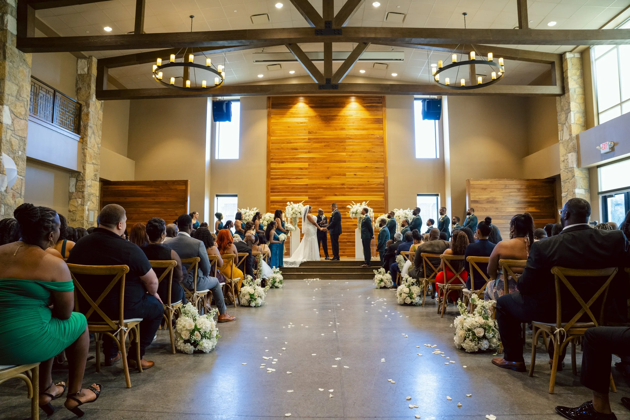 A wedding ceremony inside a modern, spacious chapel with a high ceiling, stone walls, wooden beams, and large windows. The bride and groom are at the altar exchanging vows, with guests seated on both sides decorated with white flowers and petals on t