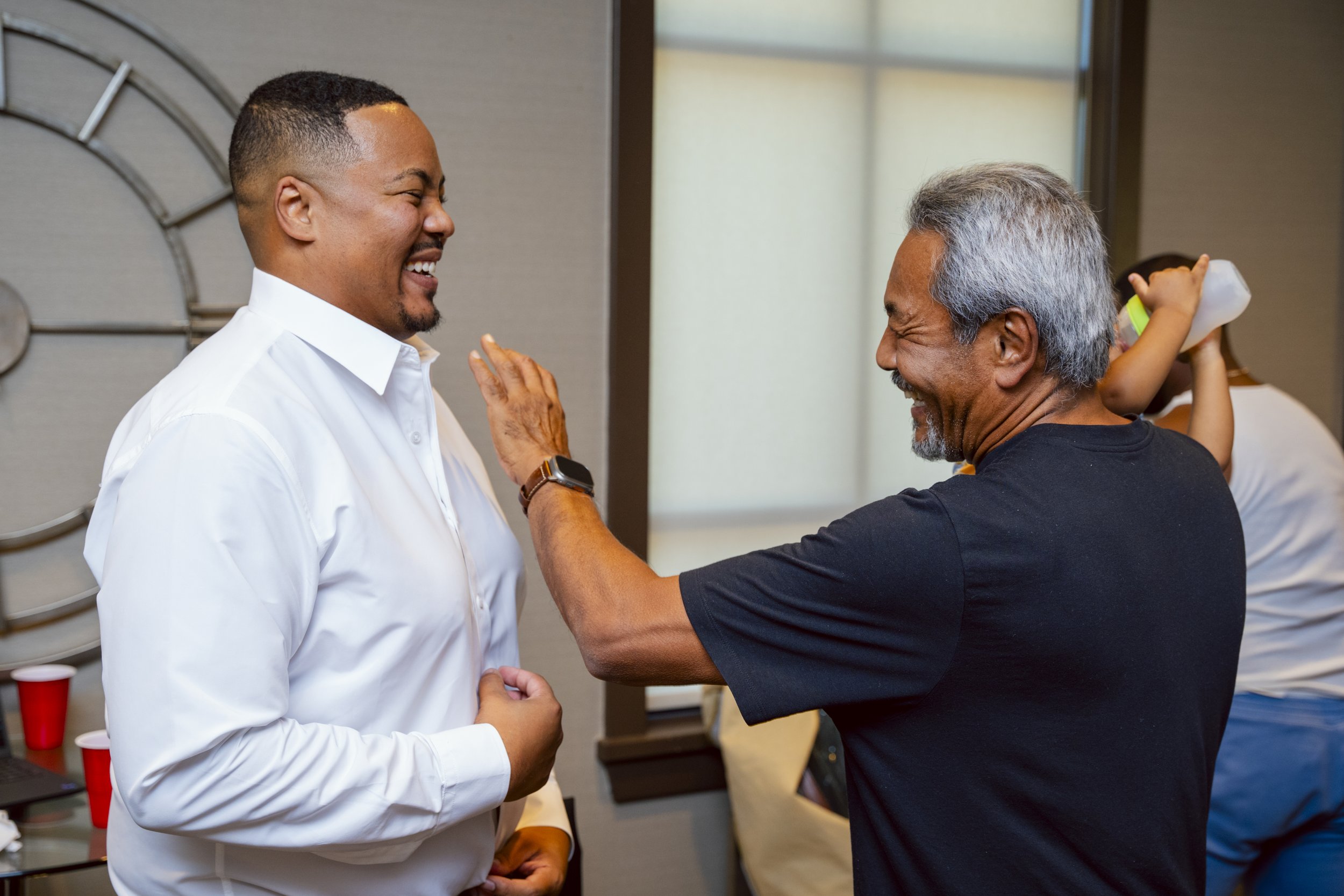 Two men are laughing and smiling at each other, one older with gray hair wearing a black t-shirt and the other younger with short dark hair wearing a white shirt, in a lively indoor setting.