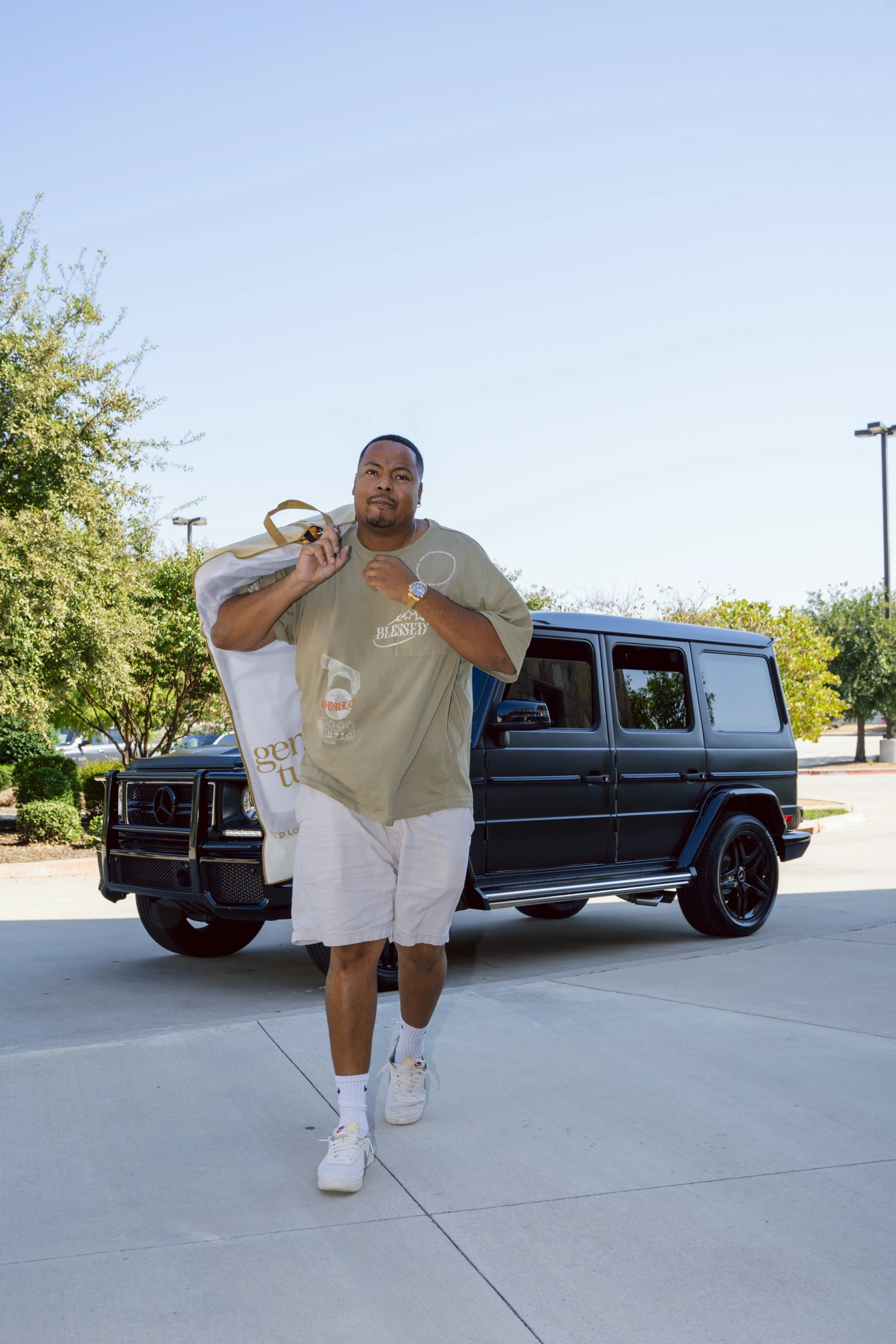 A man standing in front of a black Mercedes Benz G-Class SUV outdoors, carrying a large white shopping bag on his shoulder, wearing a beige T-shirt, white shorts, and white sneakers.