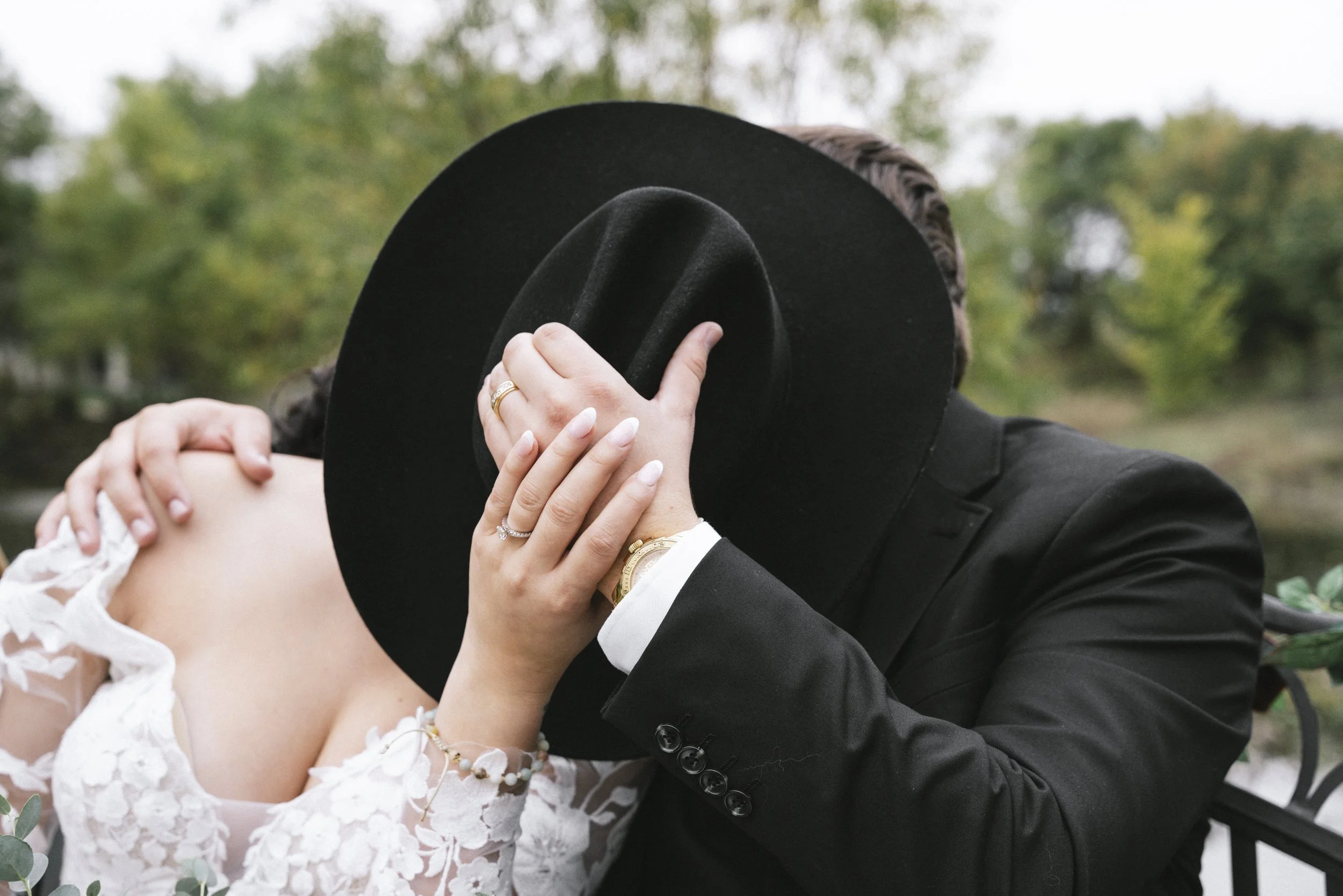 A couple sharing an intimate moment outdoors, with the man covering his face with a black hat and the woman touching his face, both in wedding attire.