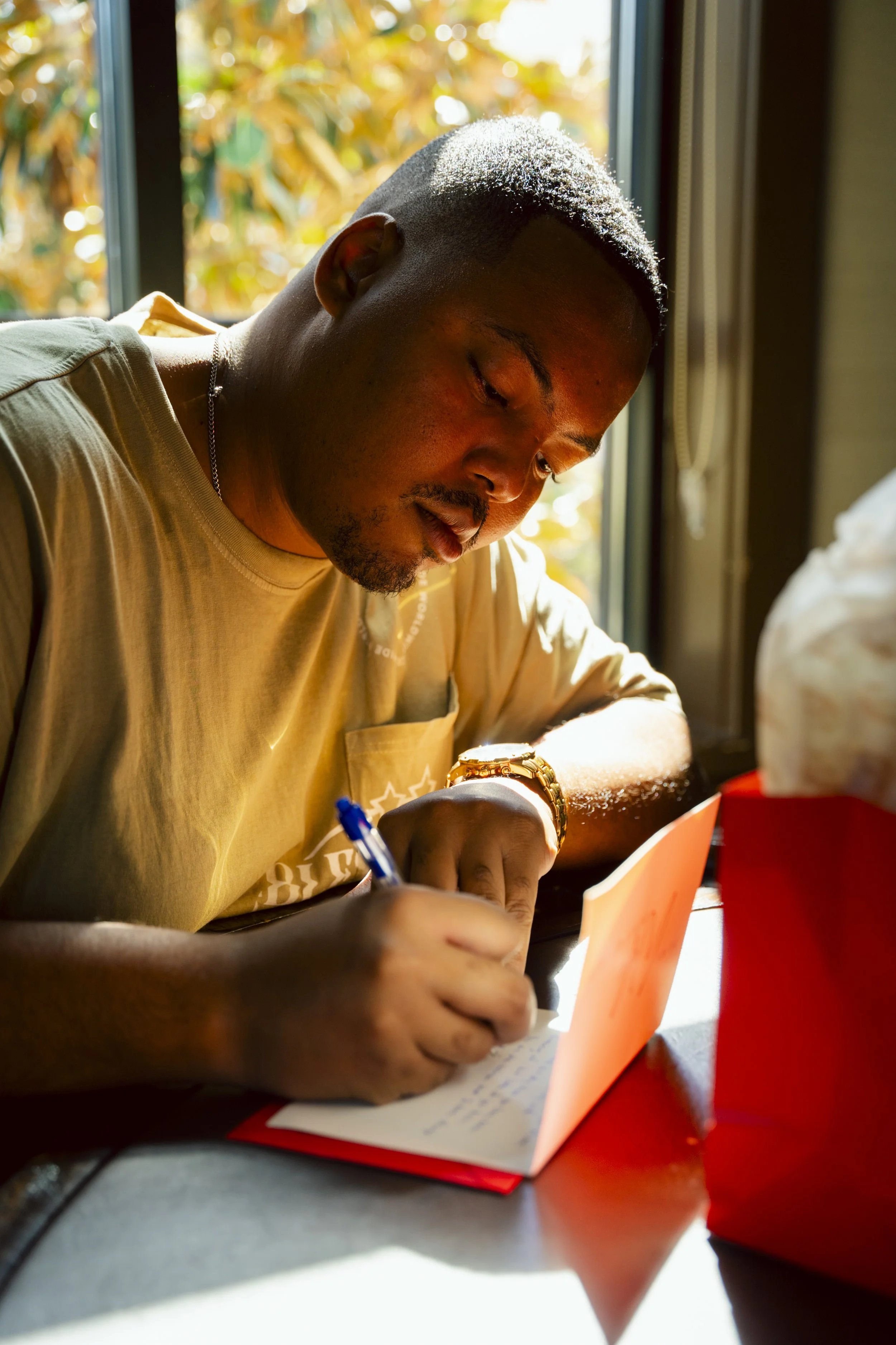 A man with short hair and a watch is writing in a notebook, sitting by a window with sunlight and autumn leaves outside.