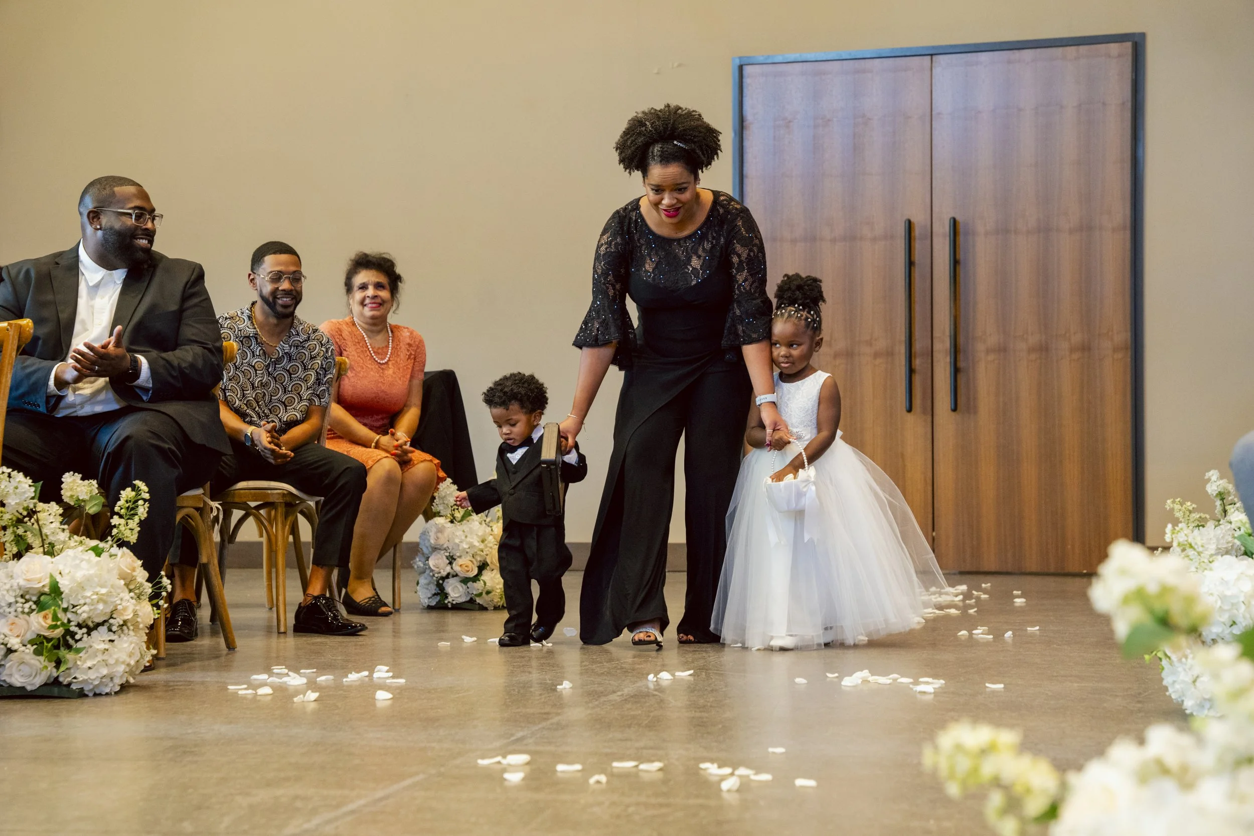 A woman with two young children walking down an aisle decorated with white flowers and flower petals, during a celebration or ceremony, with seated guests on the side watching and smiling.