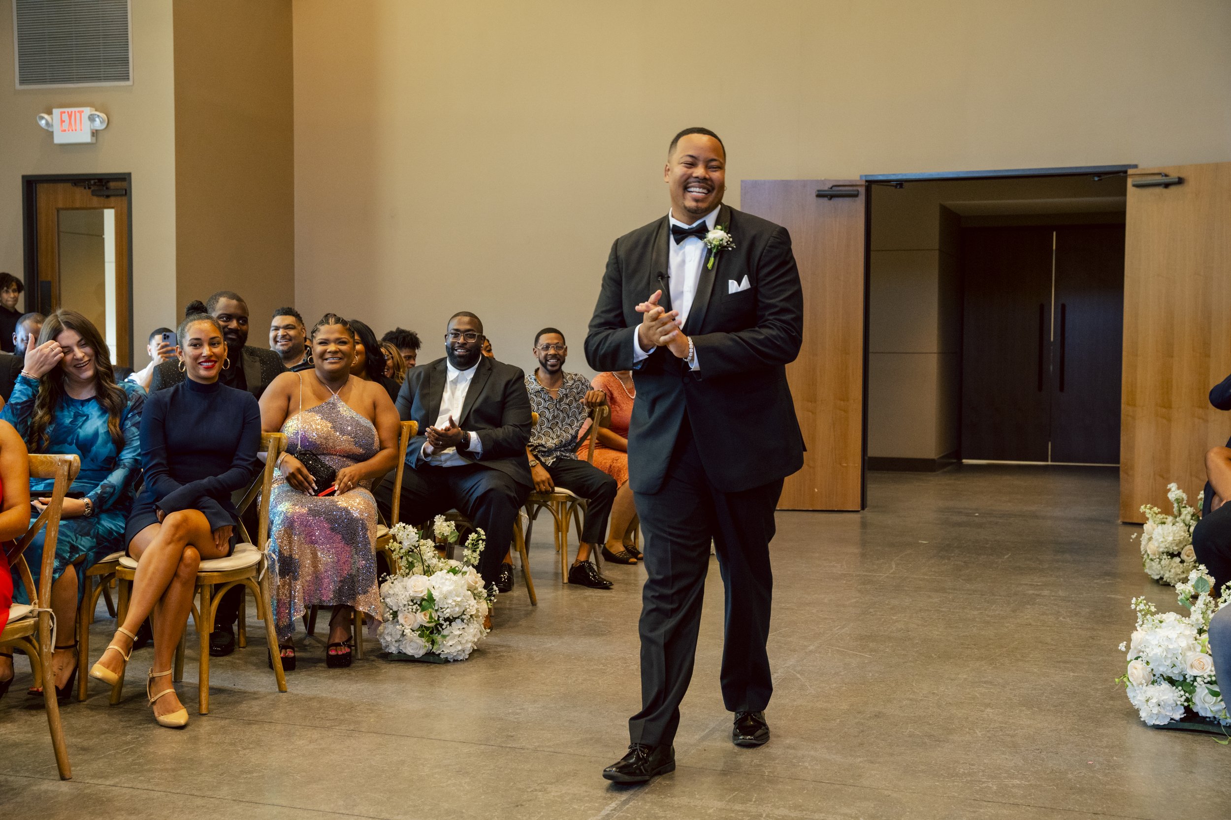 A man in a black tuxedo walking in front of seated guests at a wedding reception, smiling and clapping, with floral arrangements on the floor.