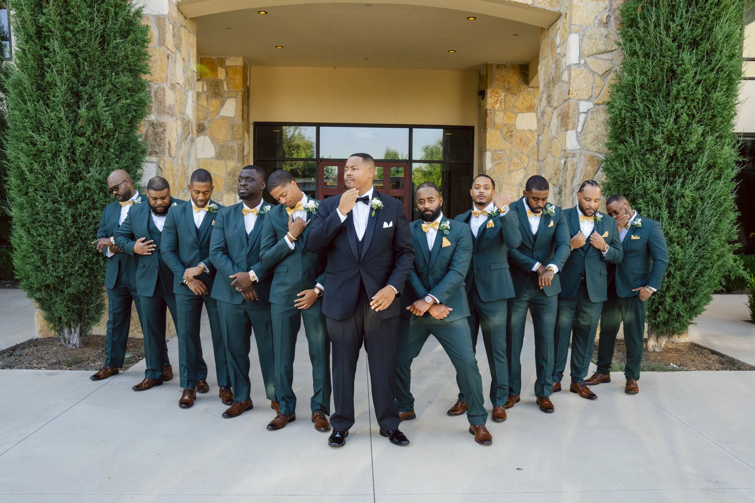 A group of twelve men dressed in matching teal suits with yellow bow ties and white shirts, standing in front of a stone building entrance with two large green trees on either side. The groom in the center wears a black suit with a white shirt and bl