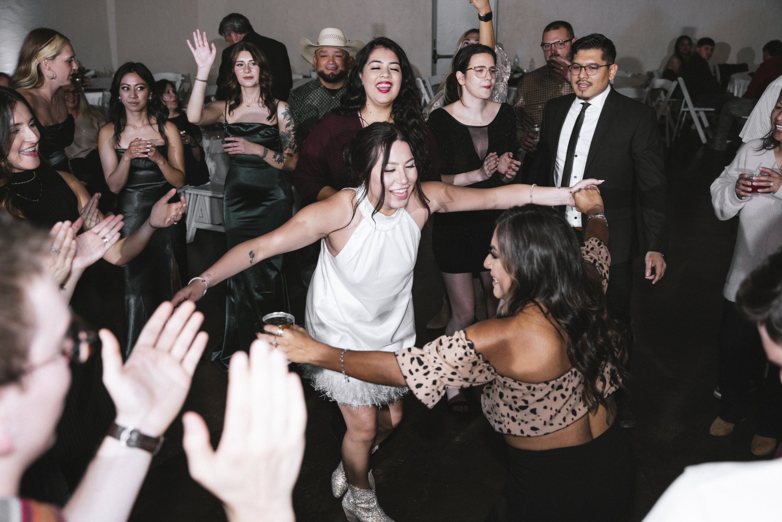 People dancing and celebrating at a party or wedding, with women in elegant dresses and a woman in a white top and feathered skirt in the center holding a drink, surrounded by friends and guests enjoying the moment.