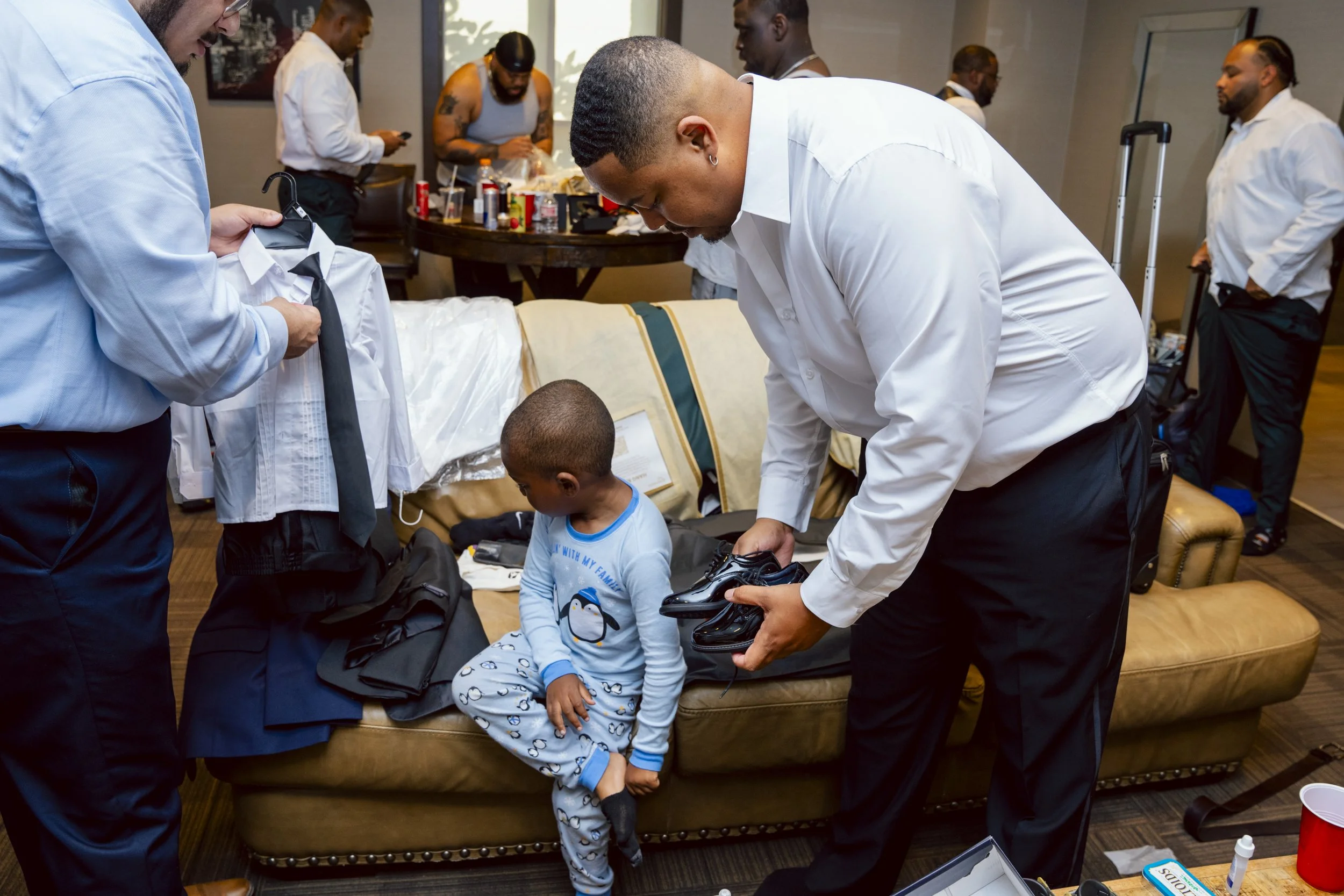 A man in a white shirt helping a young boy groom his shoes on a brown leather couch, with other men preparing or adjusting clothing in the background.
