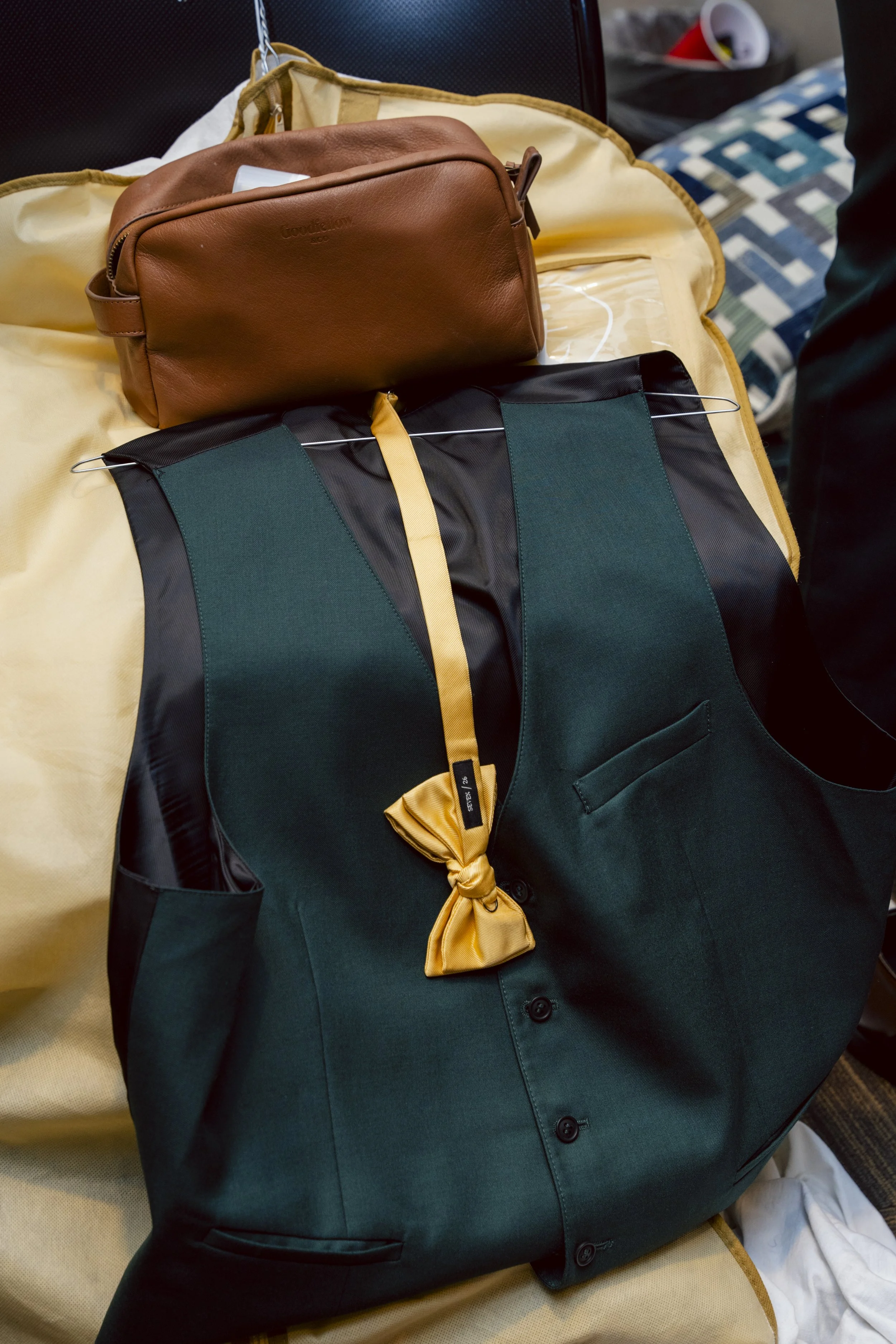 A black and green backpack with a yellow scarf tied on the handle, resting on a yellow fabric surface, with a brown leather toiletry bag on top.