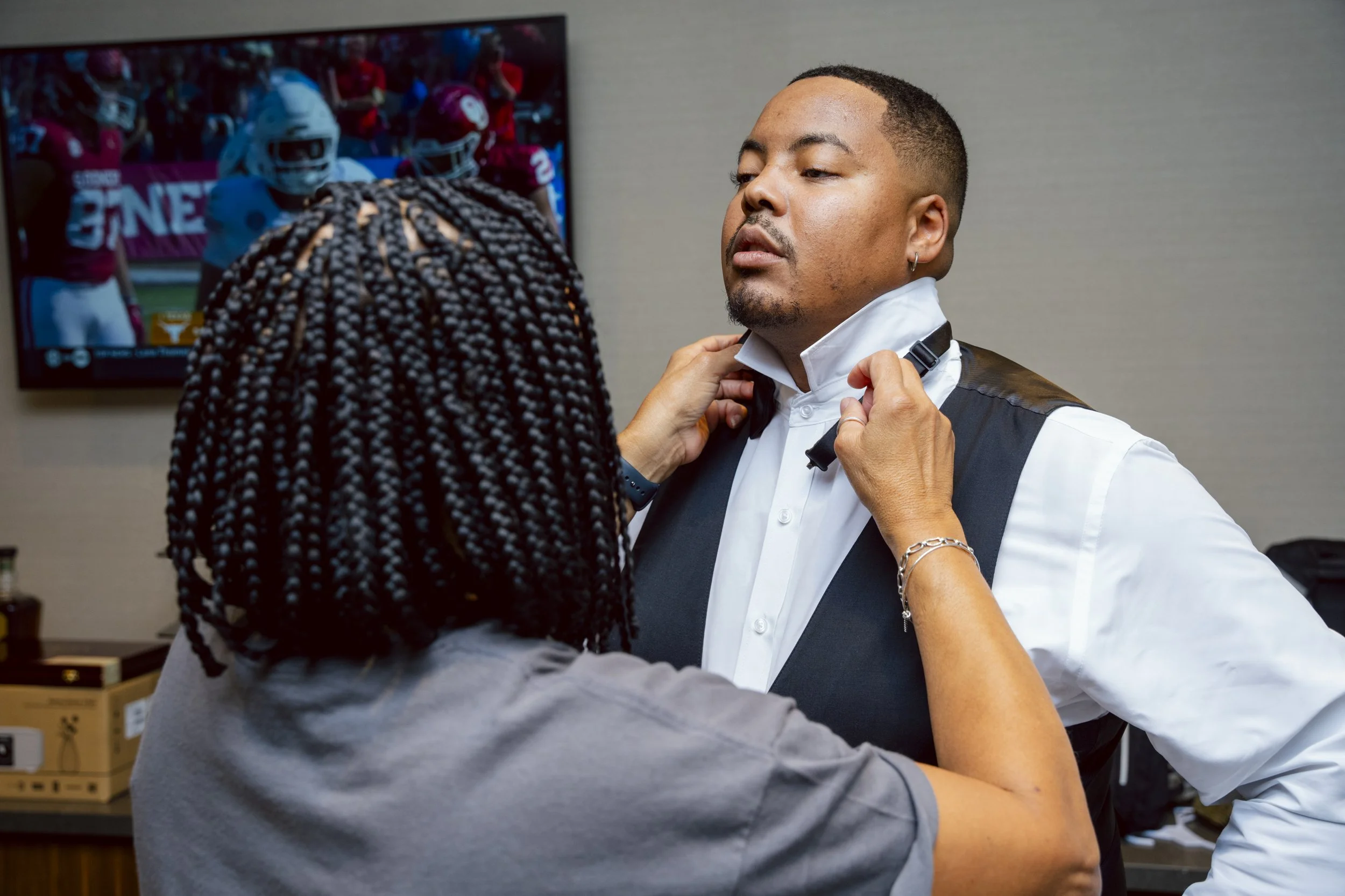 A man is being assisted by a woman in adjusting his black bow tie. The man is dressed in a white shirt and black vest, and the woman has braided hair and is wearing a gray shirt. They are in a room with a television showing a football game in the bac