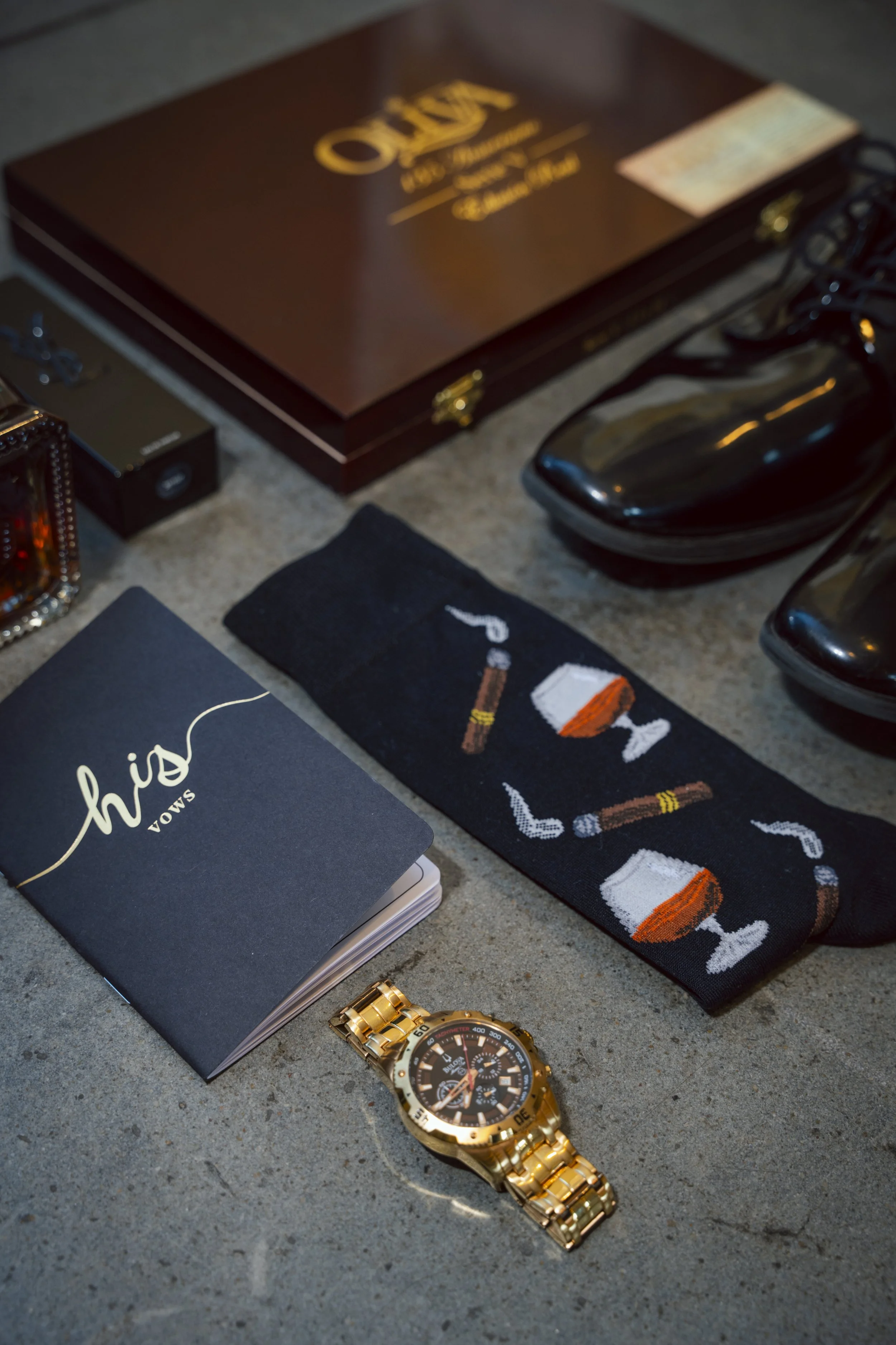 A collection of men's accessories and items on a concrete surface, including a gold wristwatch, a pair of black shoes, a dark-colored box, a black book or organizer with the word "hiso" on the cover, a pair of socks with cigar and glass printed on th