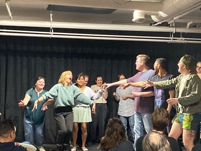 Photo of improvisers of all levels playing, one is playing Cher, pointing at Cher's Fringe Jacket, which is masterfully played by our improvisers.