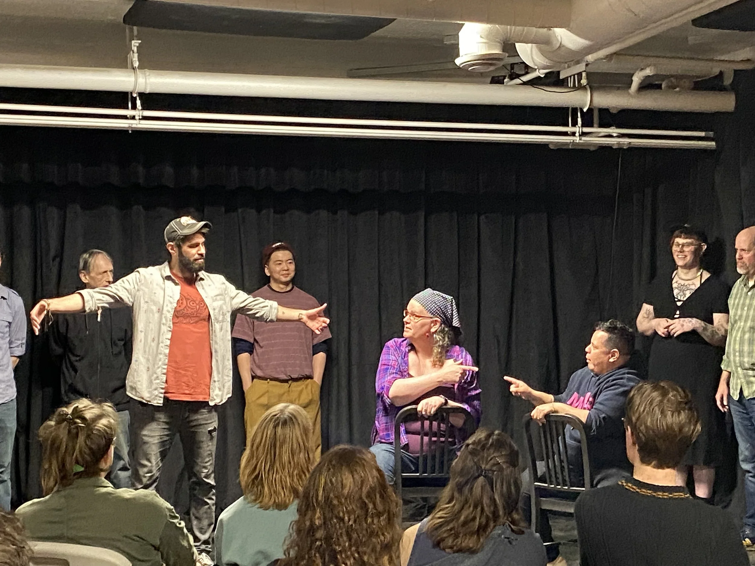 Photo of improvisers of all levels playing characters on stage, two are sitting in a chair pointing at each other and one is playing a dancing bear! 