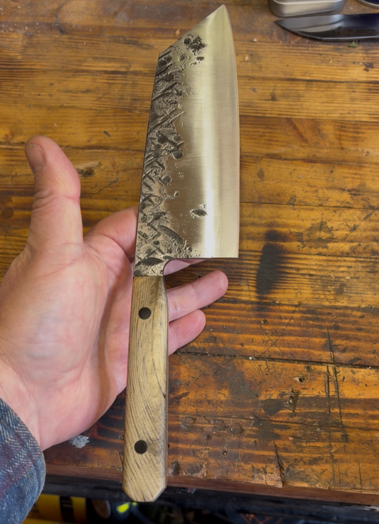 Handmade custom kitchen knife with a textured blade and light wood handle, held above a wooden workbench.