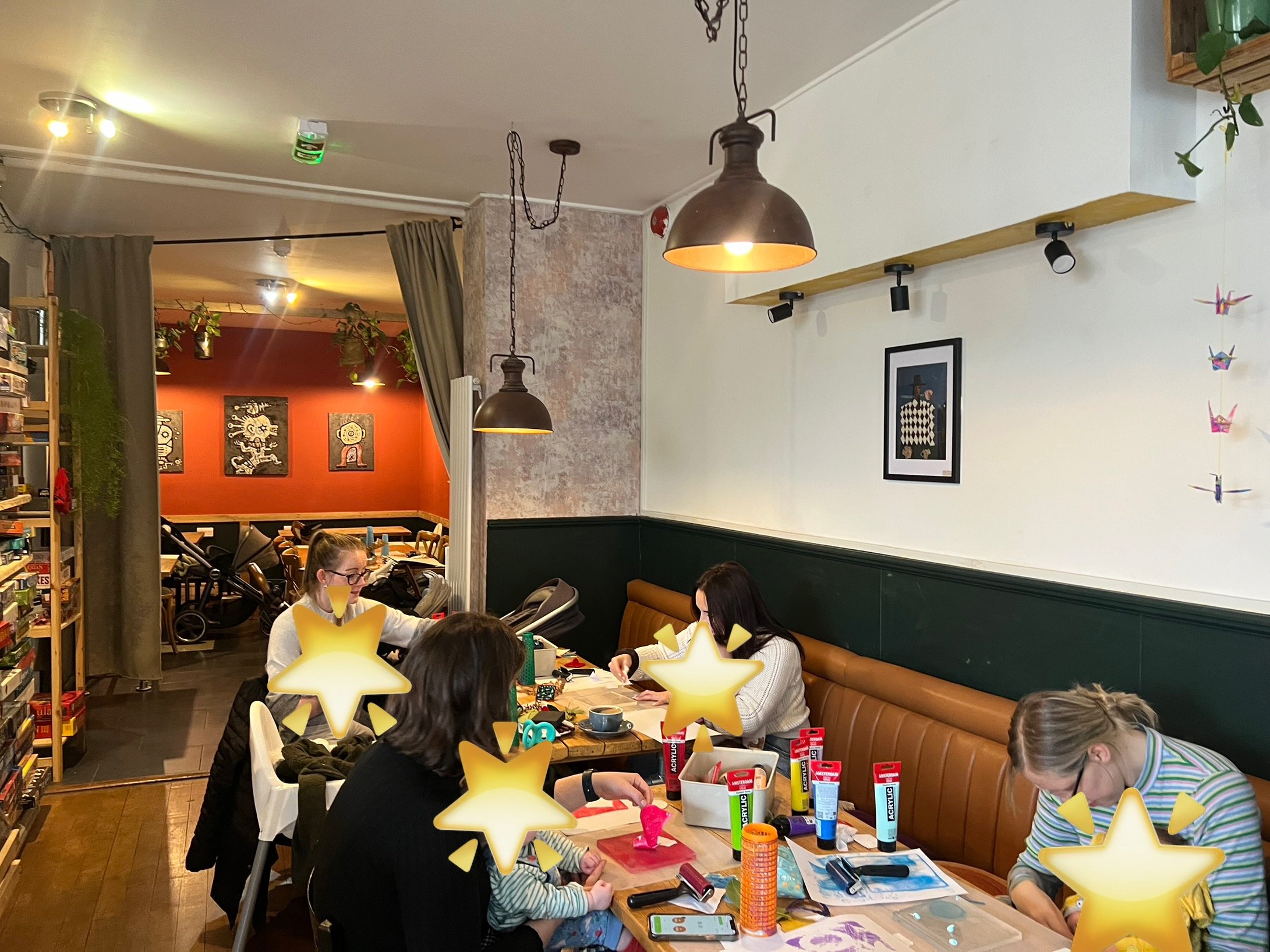 Interior of a cozy cafe with people sitting around a table doing crafts. The table is covered with art supplies including paint tubes and brushes. Shelves of board games are visible, and the walls are decorated with framed pictures and hanging lights