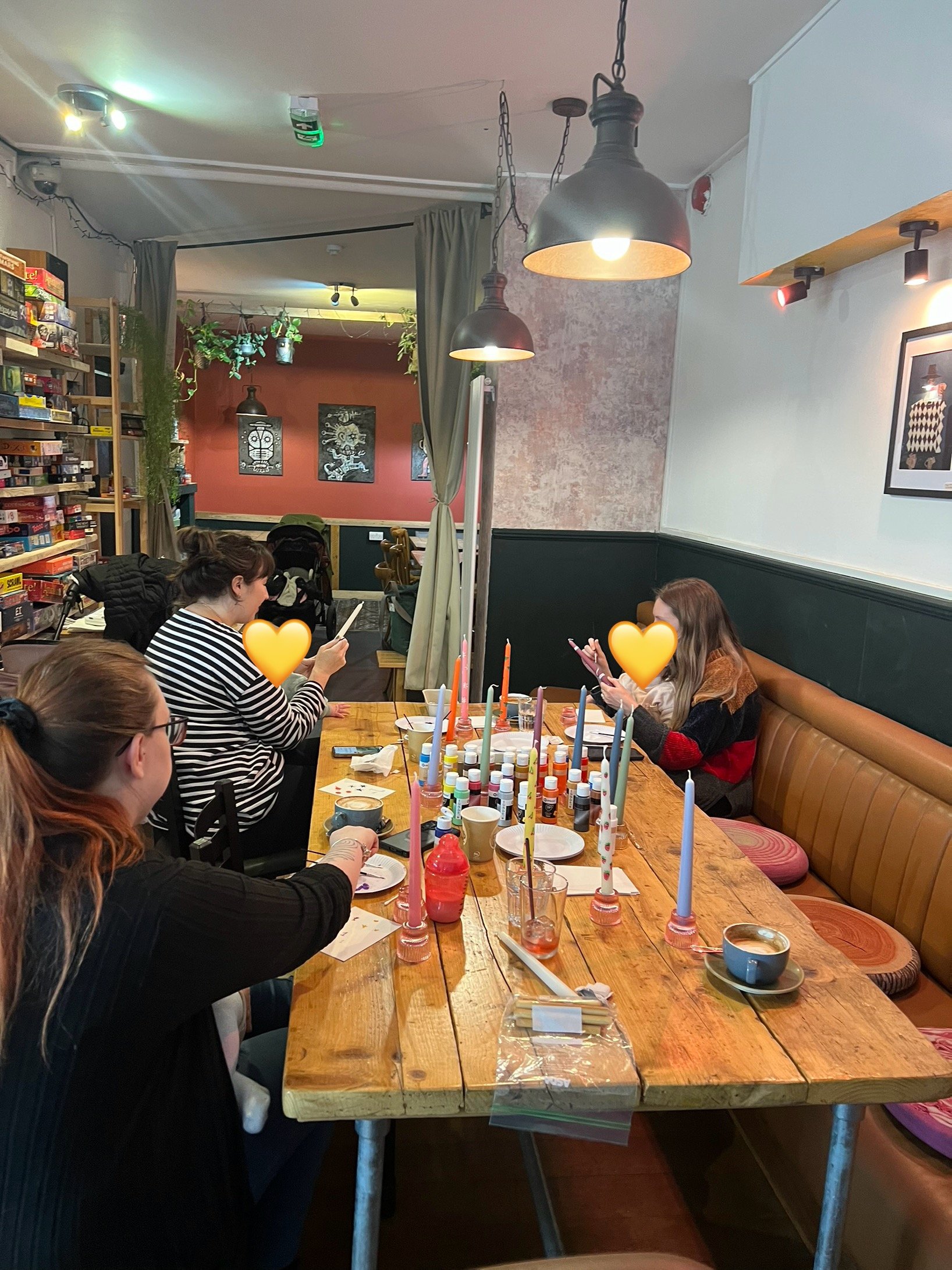 Group of people seated at a table in a cozy, board game café setting, with various colorful candles and painting supplies on the table. Shelves of board games are visible, and the ambiance is warm with hanging lights and decorative art on the walls.