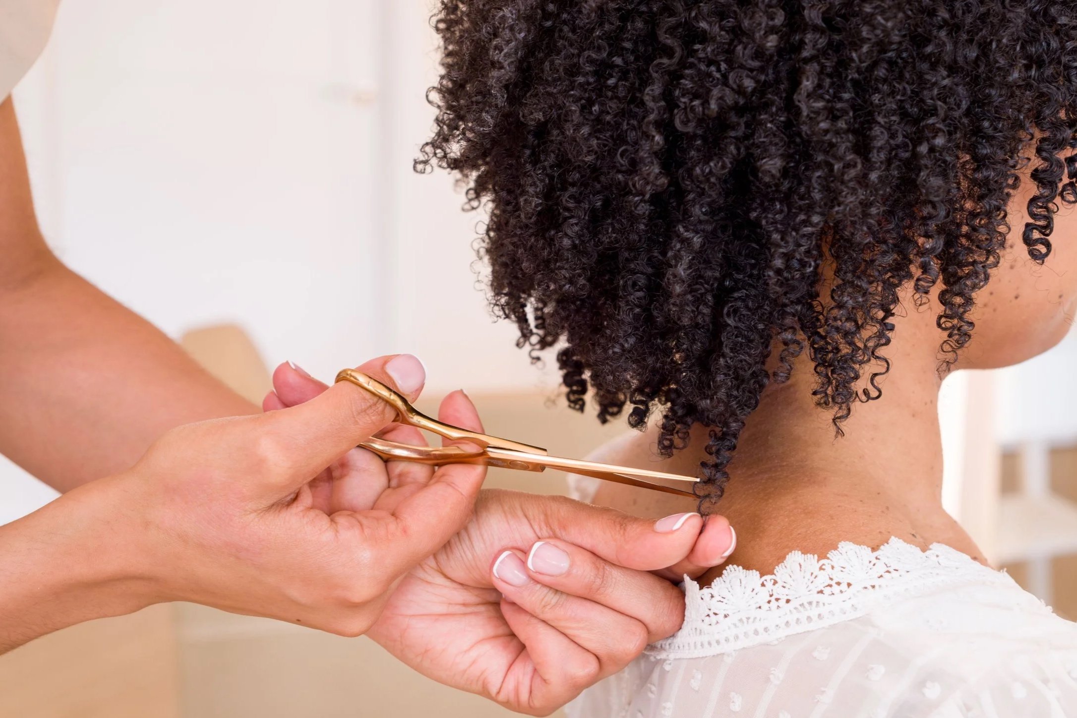 Person with curly black hair getting a haircut with scissors, while another person supports their head.