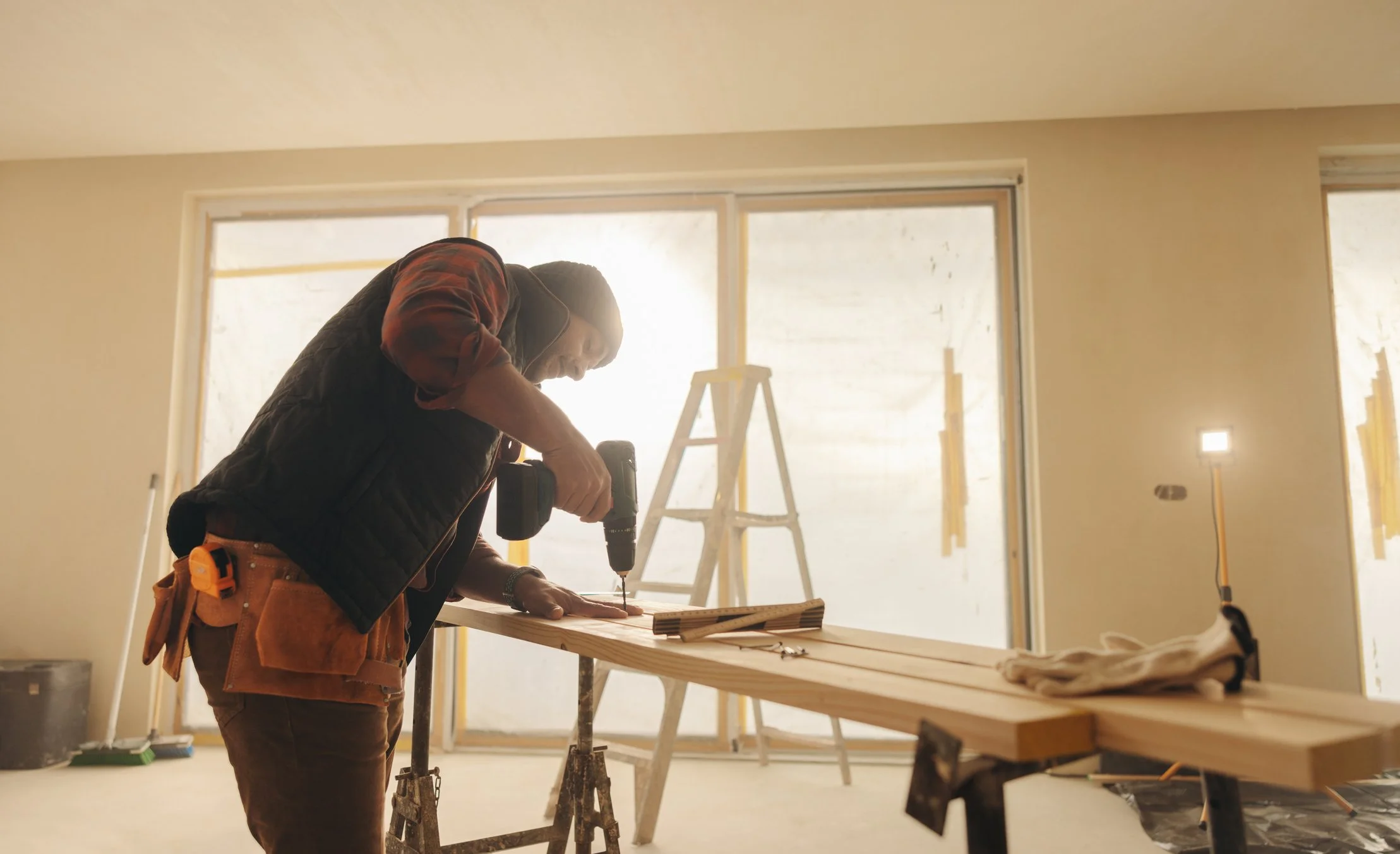 A person using a cordless drill while working on a wooden piece inside a room under construction, with a step ladder and construction tools around.